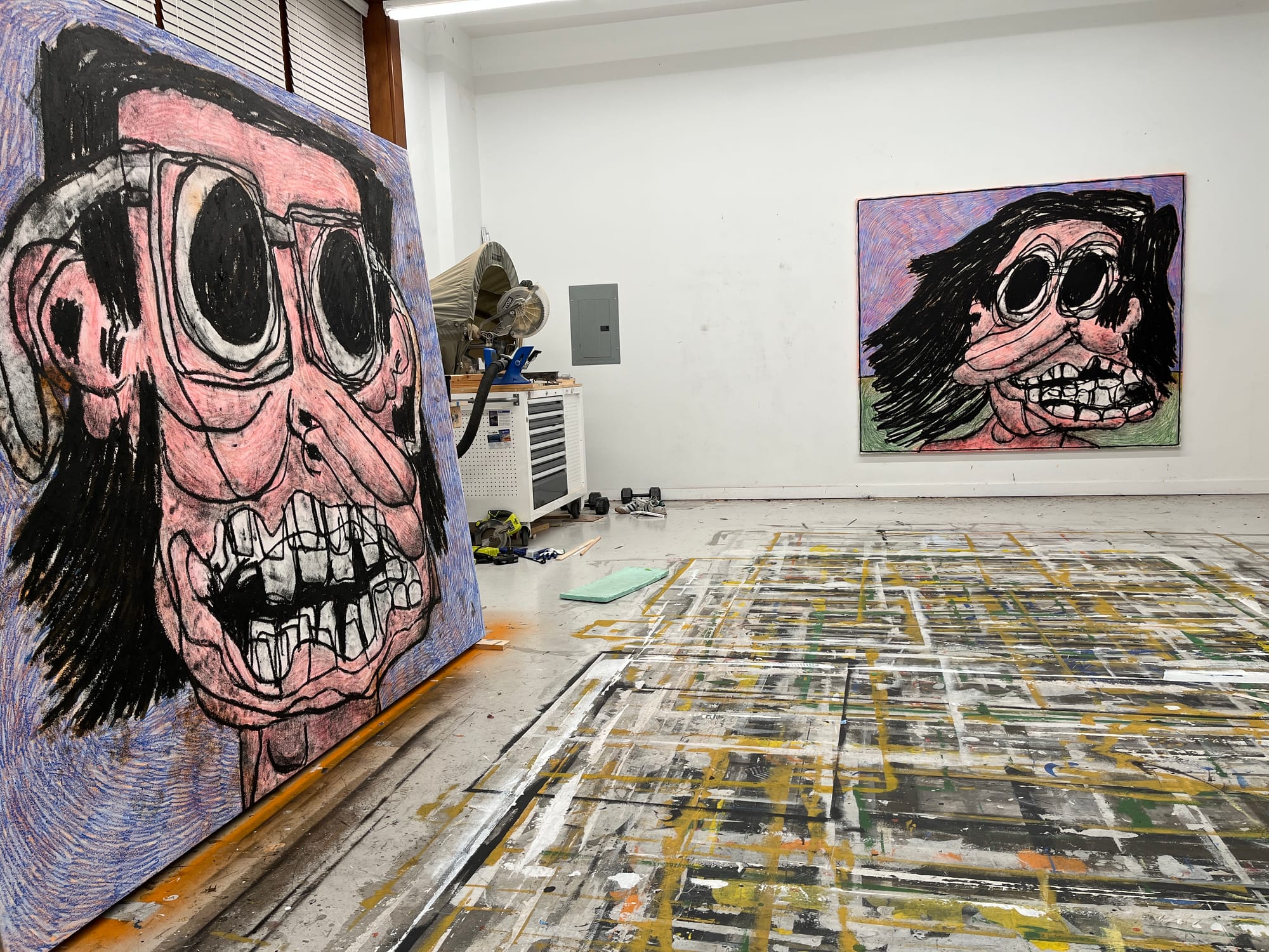 Studio view of artist Taylor A. White preparing large-scale expressionist paintings ‘Dawn, Wind’ (2023) for his upcoming exhibition at The Hole gallery, New York. Two vividly colored, cartoonish yet intense portraits with exaggerated facial features and textured backgrounds, shown in-progress amid the artist’s paint-splattered studio floor and tools. Image taken during interview with De Franco for Munchies Art Club