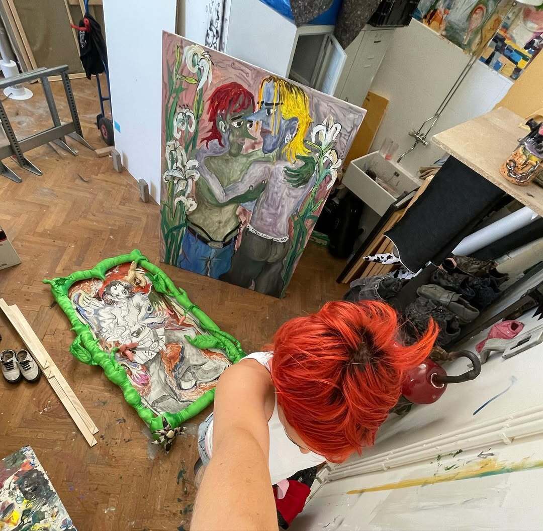 Overhead studio view of Ju Aichinger with bright red hair; large figurative painting leaning against the wall and a neon-green framed relief on the floor; tools, shoes, and materials scattered—Academy of Fine Arts Vienna studio.