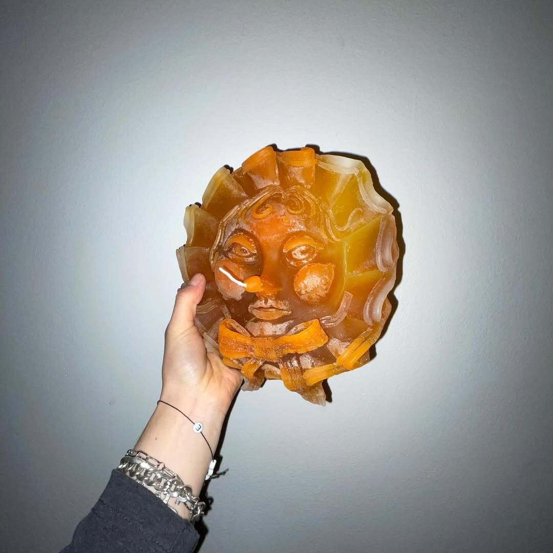 Ju Aichinger “Candle Faces” work-in-progress—hand holding an amber, sun-faced cast object against a gray wall; intimate studio snapshot of the series.