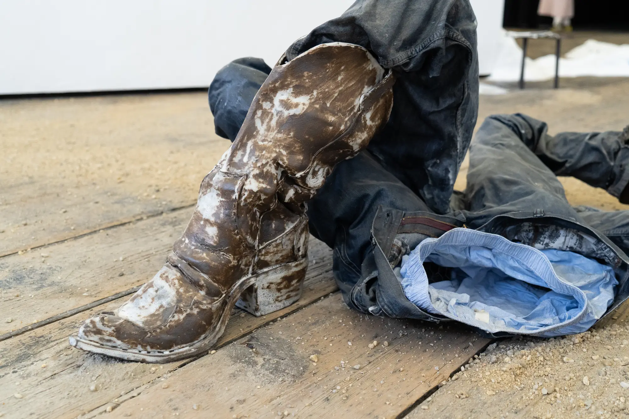 Detail from Ju Aichinger’s installation “to be or not to be (butch)”—mud-smeared boots beside crumpled jeans and blue shirt on a timber floor; queer presence/absence; shown at the Academy of Fine Arts Vienna, Semperdepot