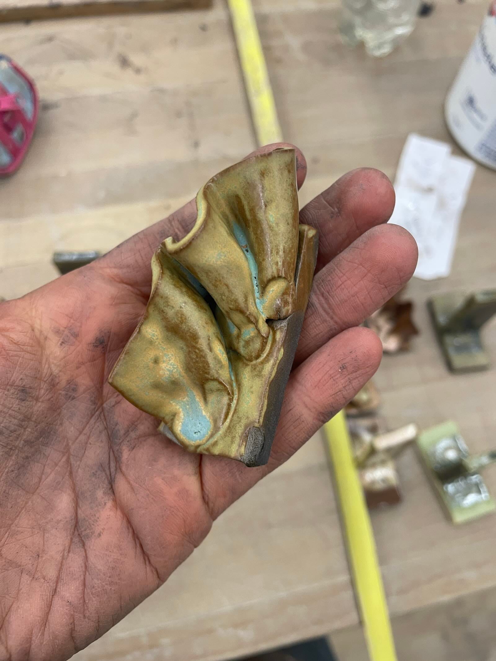 Ju Aichinger studio WIP—close-up of a hand holding a small folded piece of green-gold glazed ceramic with blue accents; tools and test tiles blurred on the worktable.