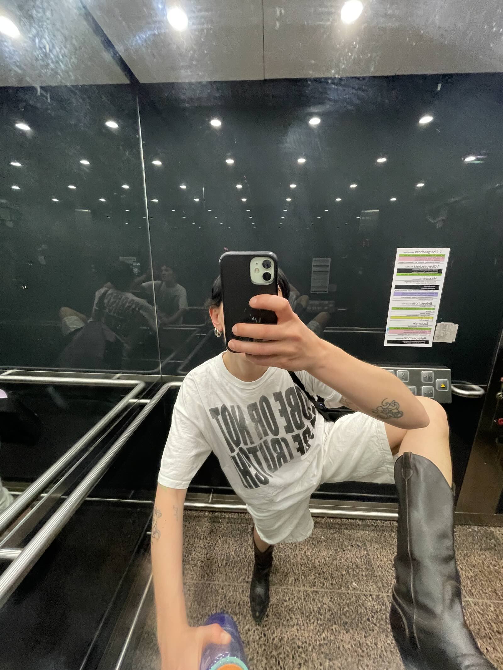 Ju Aichinger elevator mirror self-portrait—white oversized T-shirt, one leg raised in a black boot, phone covering face; closing image for Munchies Art Club Artist Spotlight; Vienna-based artist working across ceramics, costume, and performance.