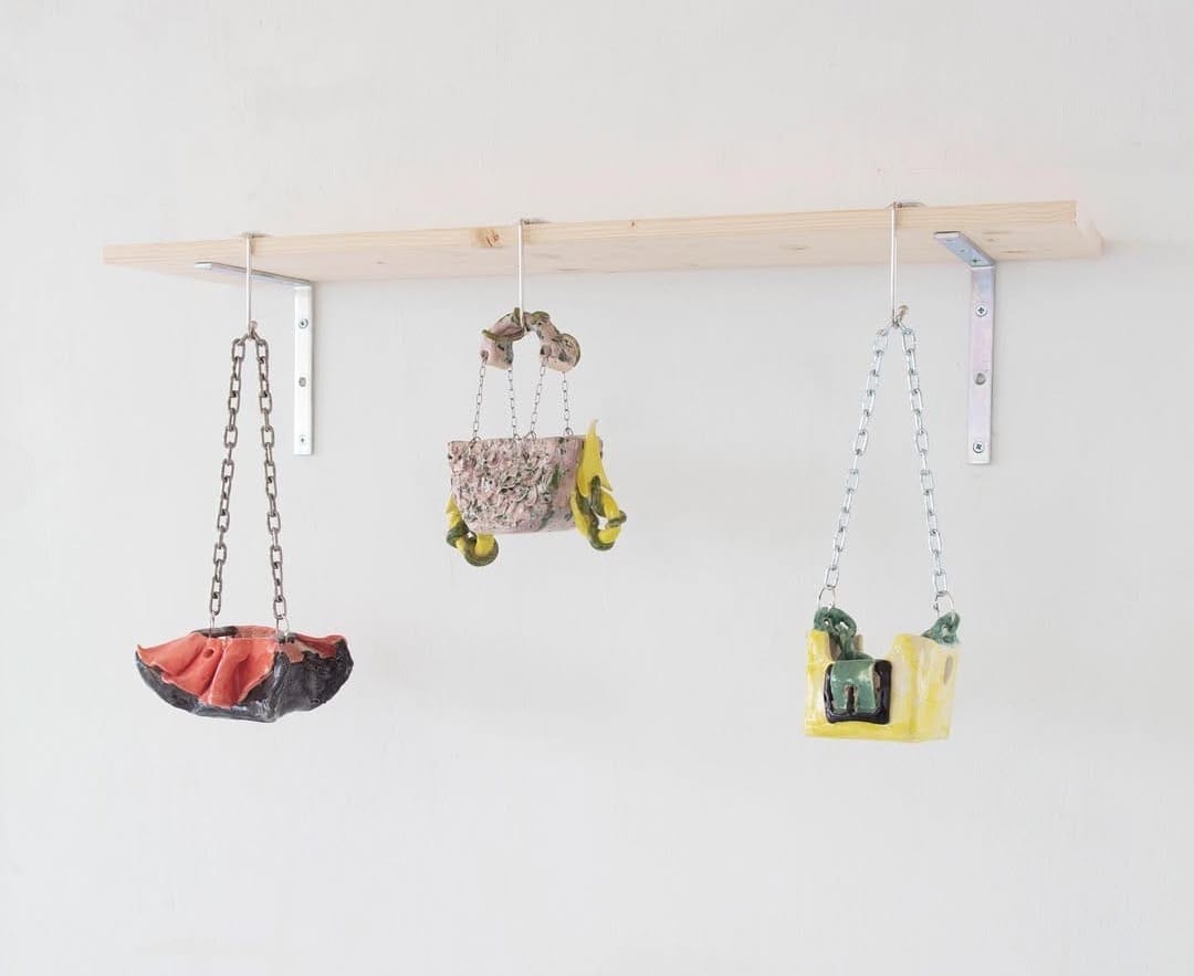 Three small glazed ceramic sculptures resembling handbags/baskets, each hanging by metal chains from a wooden shelf on a white wall—exhibition view by Ju Aichinger at the_dessous.
