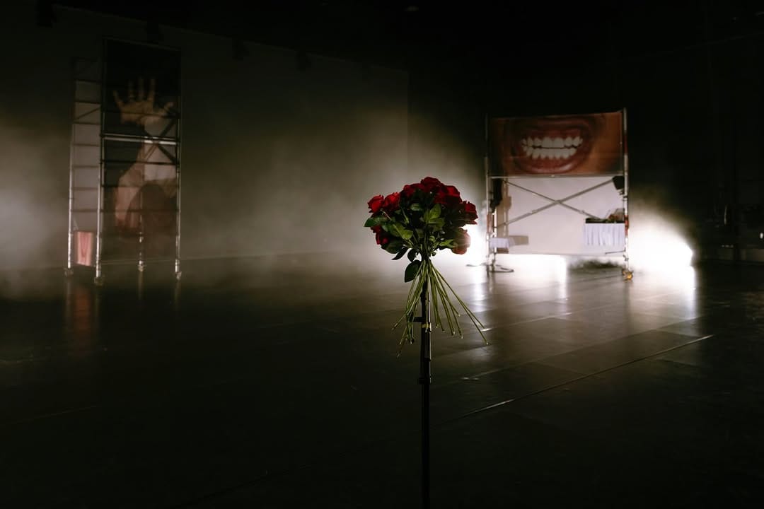 Performance “hold yr ache 2 my ache” — two rolling scaffold towers wrapped in translucent prints of face/mouth/hands on a dim, hazy stage; bouquet at base. Stage design & costume by Ju Aichinger with Val Holfeld; photo Marcella Ruiz Cruz.