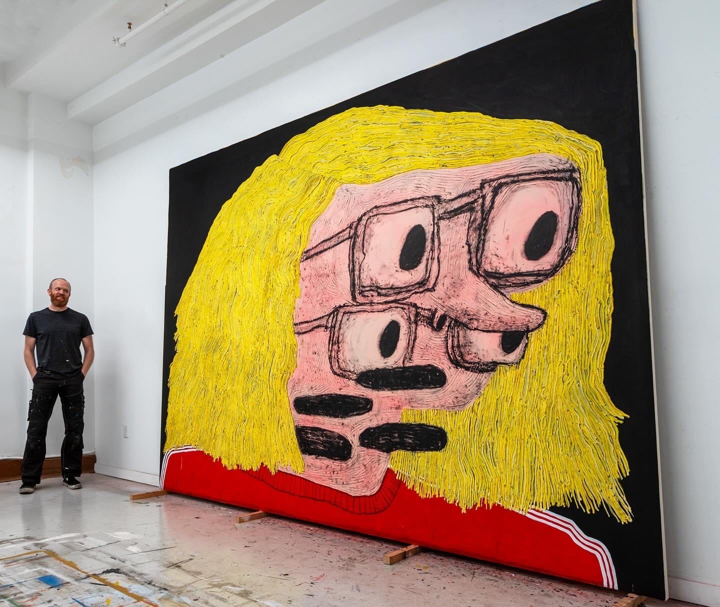 Artist Taylor A. White standing in his studio beside a large-scale painting titled Day Sight. The work shows a distorted pink face with four oversized eyes behind thick black glasses, surrounded by vibrant yellow hair and set against a black background. The figure wears a red sweater. The floor is splattered with paint and studio markings.