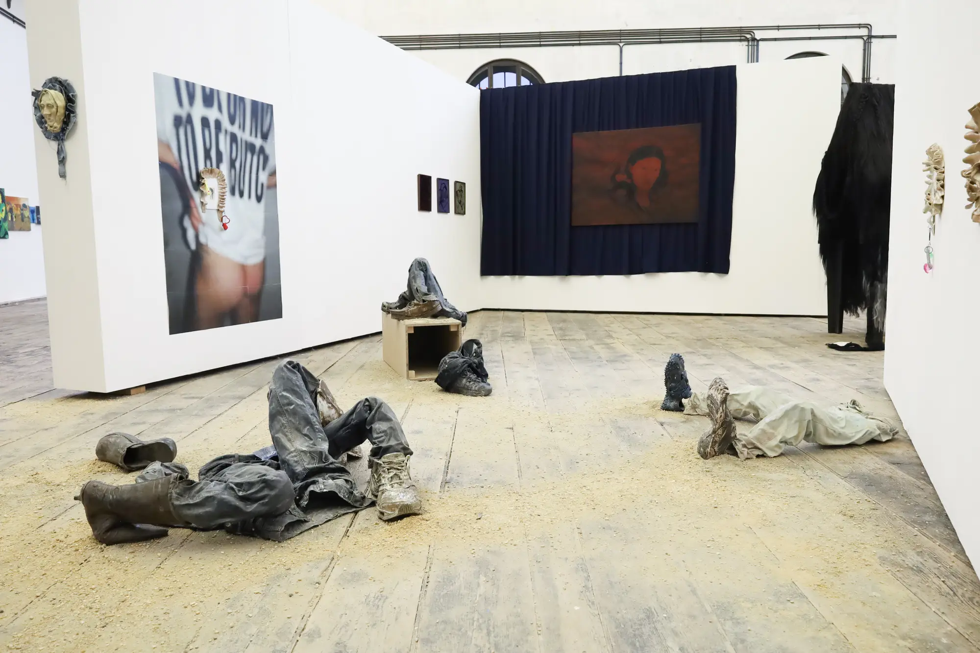 Installation view of Ju Aichinger’s “to be or not to be (butch)” at Semperdepot, Vienna—mud-smeared boots, jeans and cast garment forms spread across a wooden gallery floor; photos and works on surrounding walls.