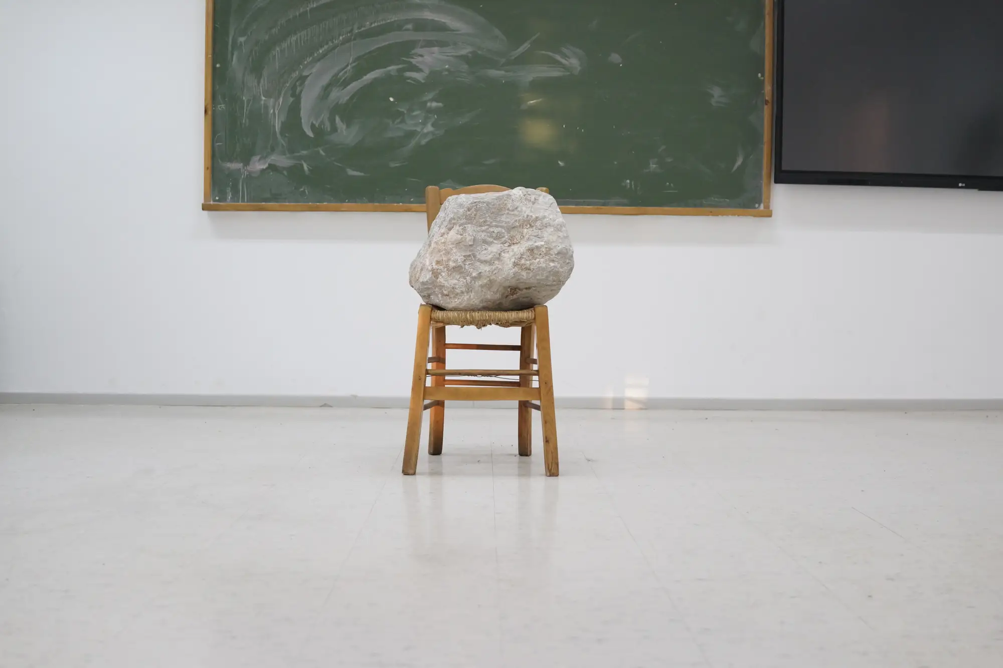 Installation by Jannis Kounellis at Hydra School Projects 2025, Lithos Lethe. A stone rests on a wooden chair in front of a green chalkboard inside the former Hydra school, connecting Arte Povera with the traces of the classroom setting.