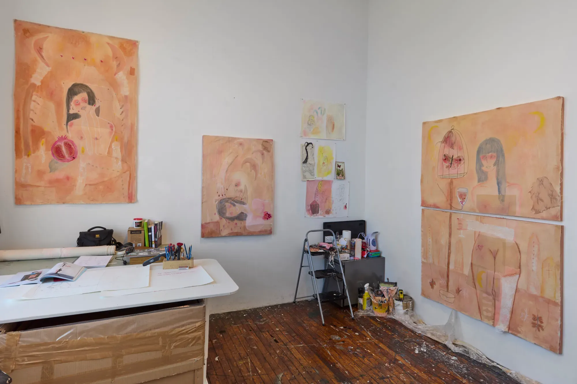 Studio view of Ailyn Lee with large skin-toned diptych (female figure, cage, moons) on right, smaller canvases and pinned sketches on left, worktable with papers and tools, paint-splattered wooden floor—new works in progress.