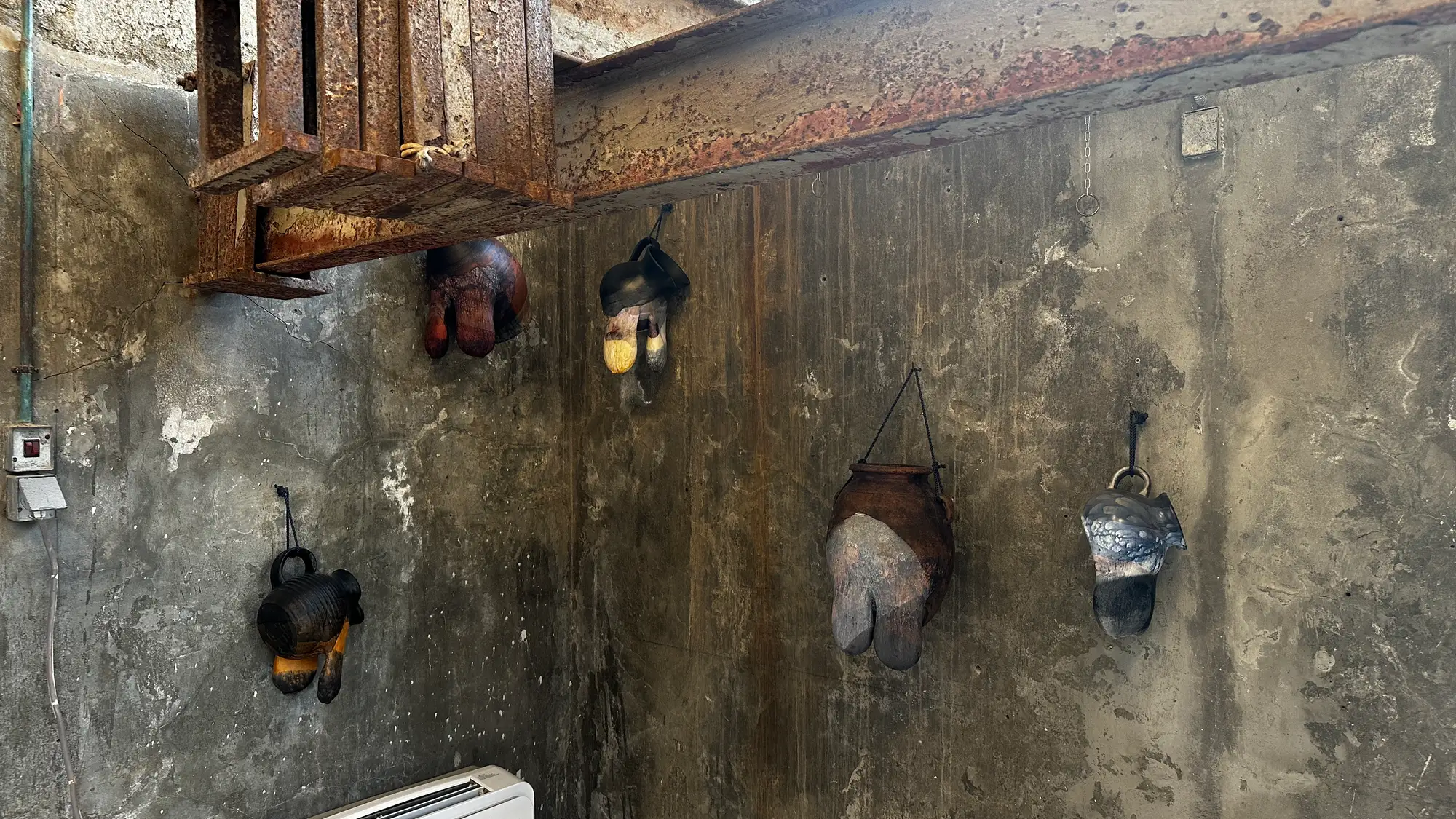 Rust-streaked interior wall at DESTE Slaughterhouse, Hydra, with five wall-hung vessel sculptures by Andra Ursuța—breast/udder-like bronze jugs with polychrome, ceramic-style patina—from “Apocalypse Now and Then,” 2025.