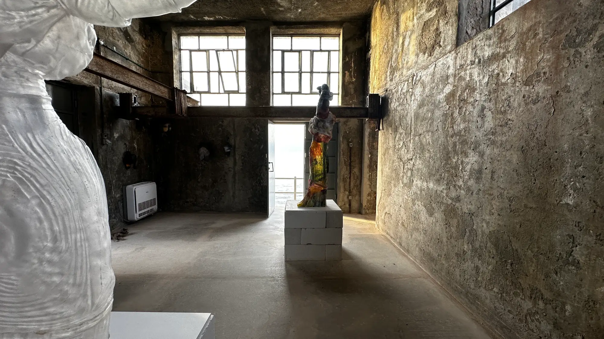 DESTE Foundation Project Space (Slaughterhouse), Hydra—installation view of Andra Ursuța’s “Apocalypse Now and Then,” 2025: translucent glass torso and colorful mixed-media figure on block plinths, rough concrete walls, sea visible through open doorway.