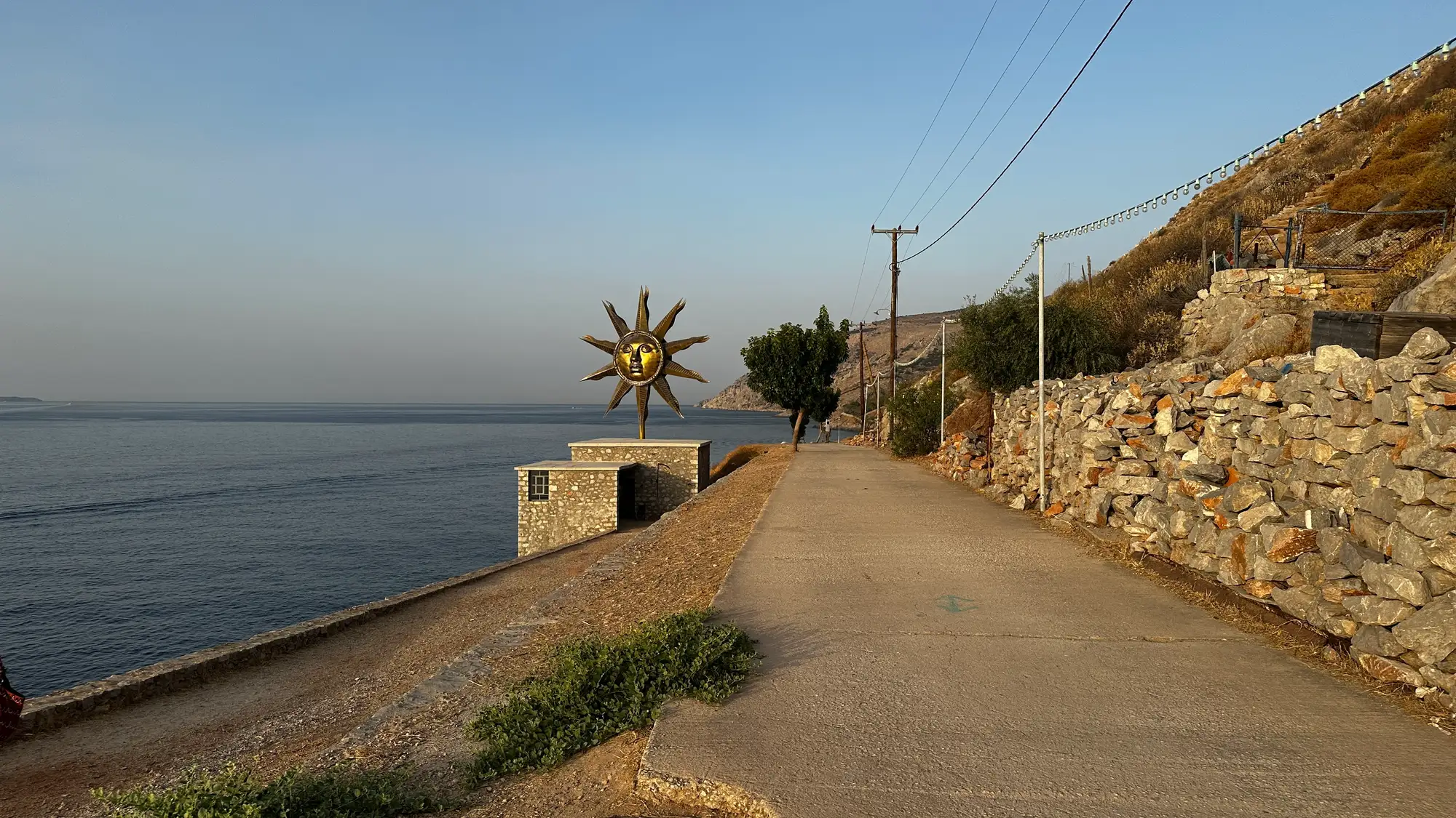 Path from Hydra port to DESTE Foundation Project Space (Slaughterhouse) with Jeff Koons’s “Apollo” wind spinner beside the sea, stone walls and power lines, late July evening on Hydra Island, Greece.
