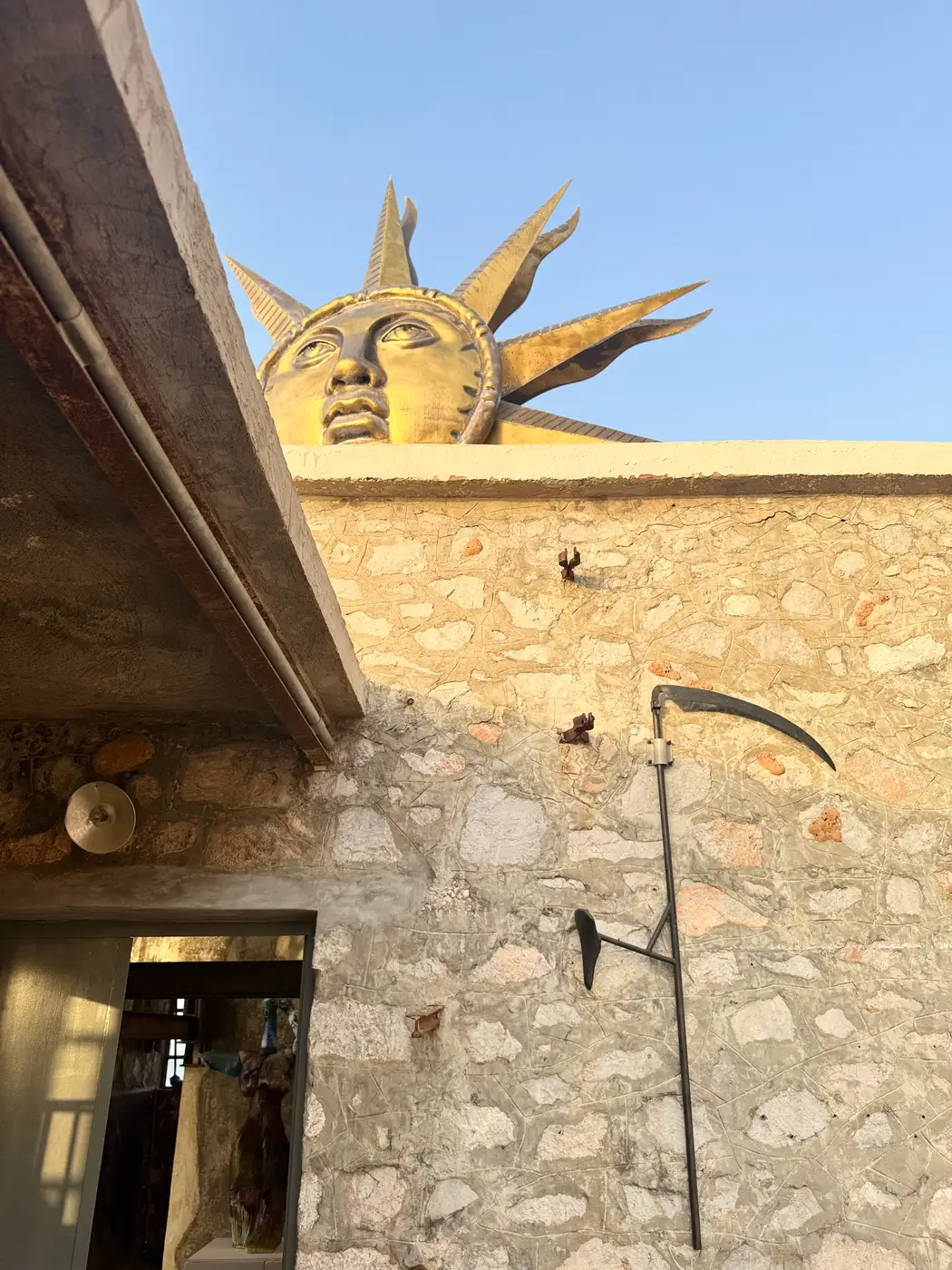 Entrance of DESTE Foundation Project Space (Slaughterhouse), Hydra—Jeff Koons’s Apollo wind spinner above stone wall, Andra Ursuța’s scythe-like wall piece at right, glass sculpture visible through doorway, 2025.