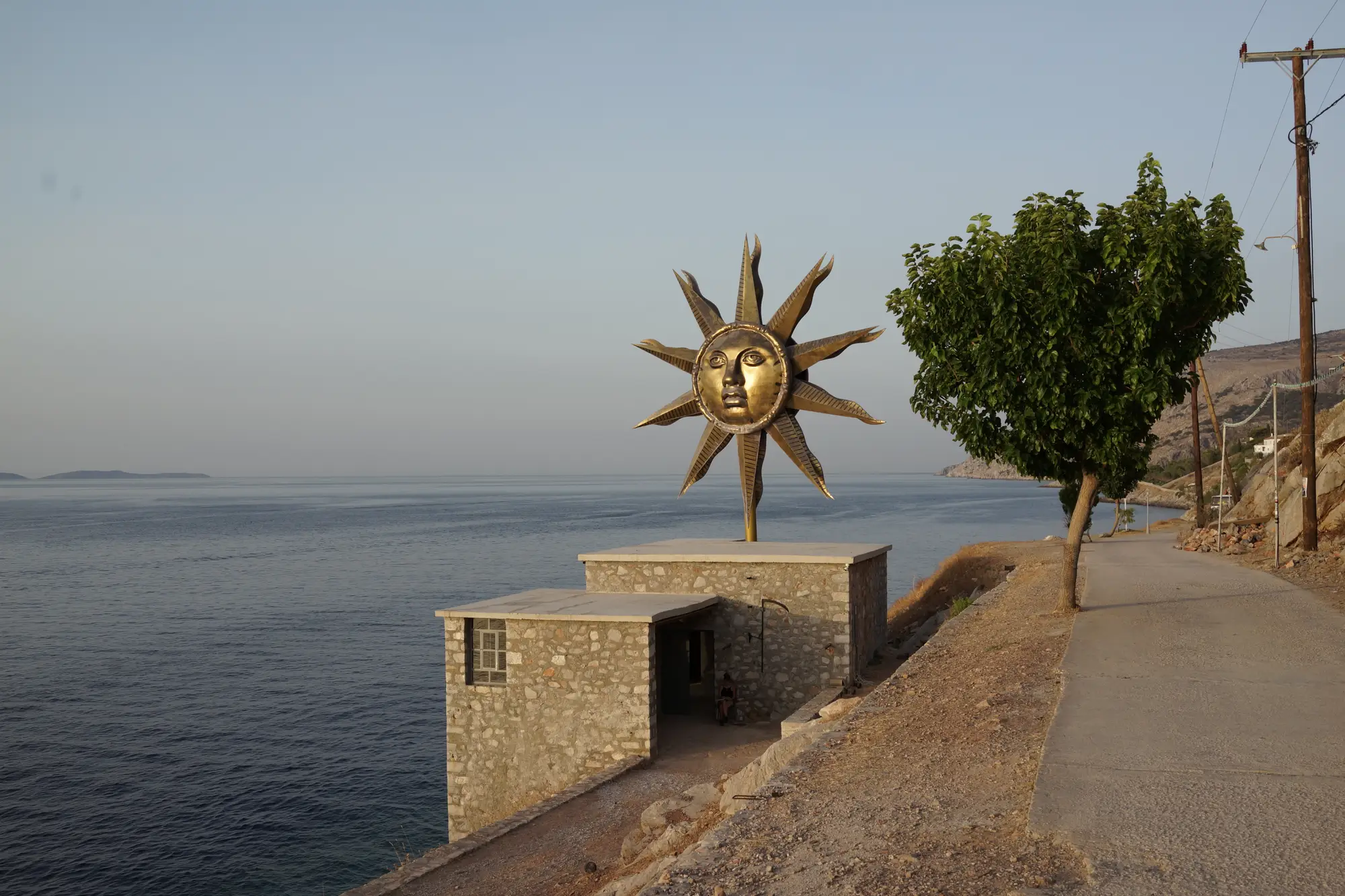 DESTE Foundation Project Space (Slaughterhouse), Hydra Island, Greece—coastal path beside stone building topped by Jeff Koons’s “Apollo” sun sculpture, evening light over the Saronic Gulf, farewell view after Andra Ursuța’s 2025 exhibition.
