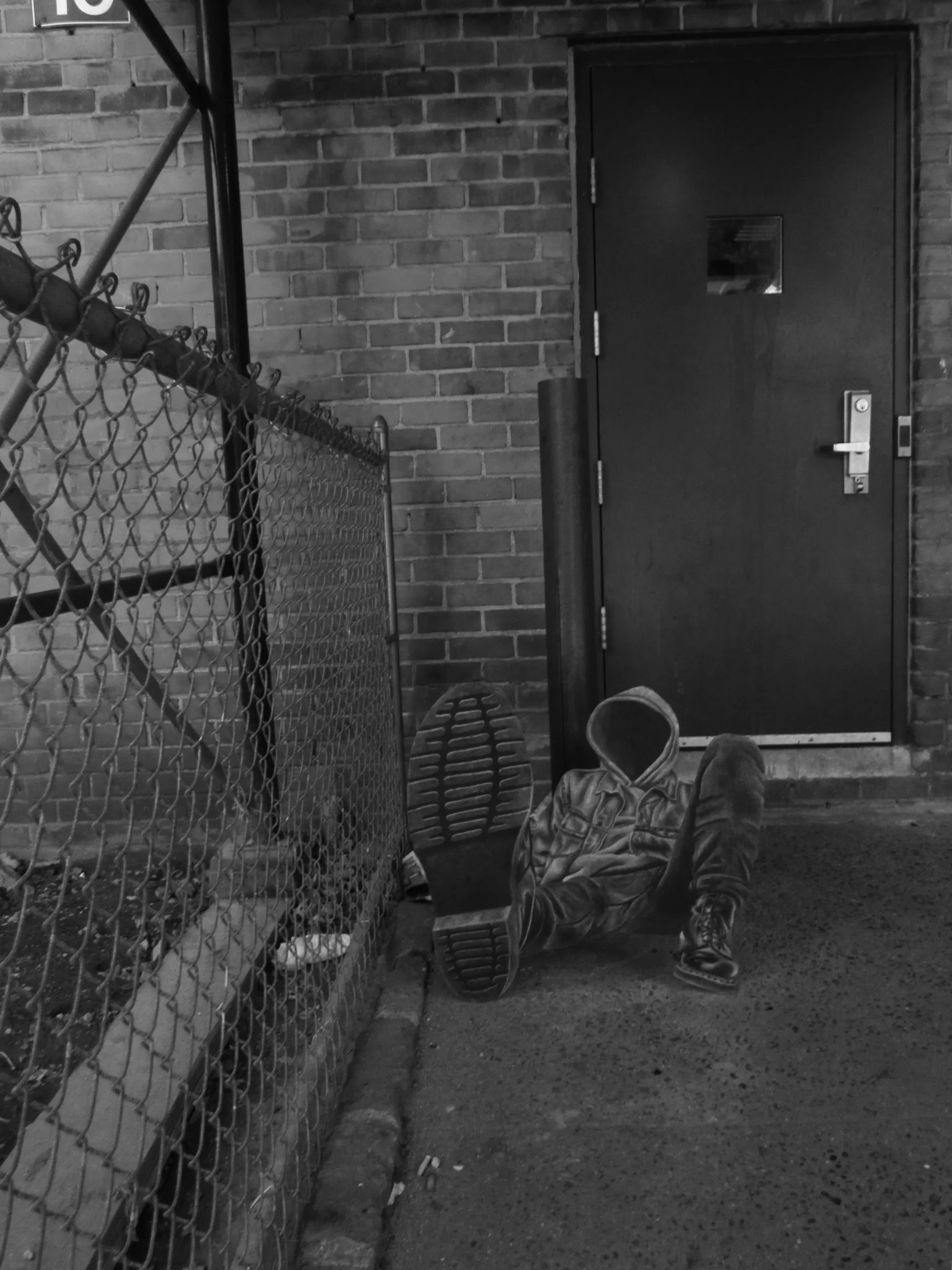 Daniel Castro black-and-white scene with hooded-figure sculpture slumped near a brick doorway and chain-link fence.