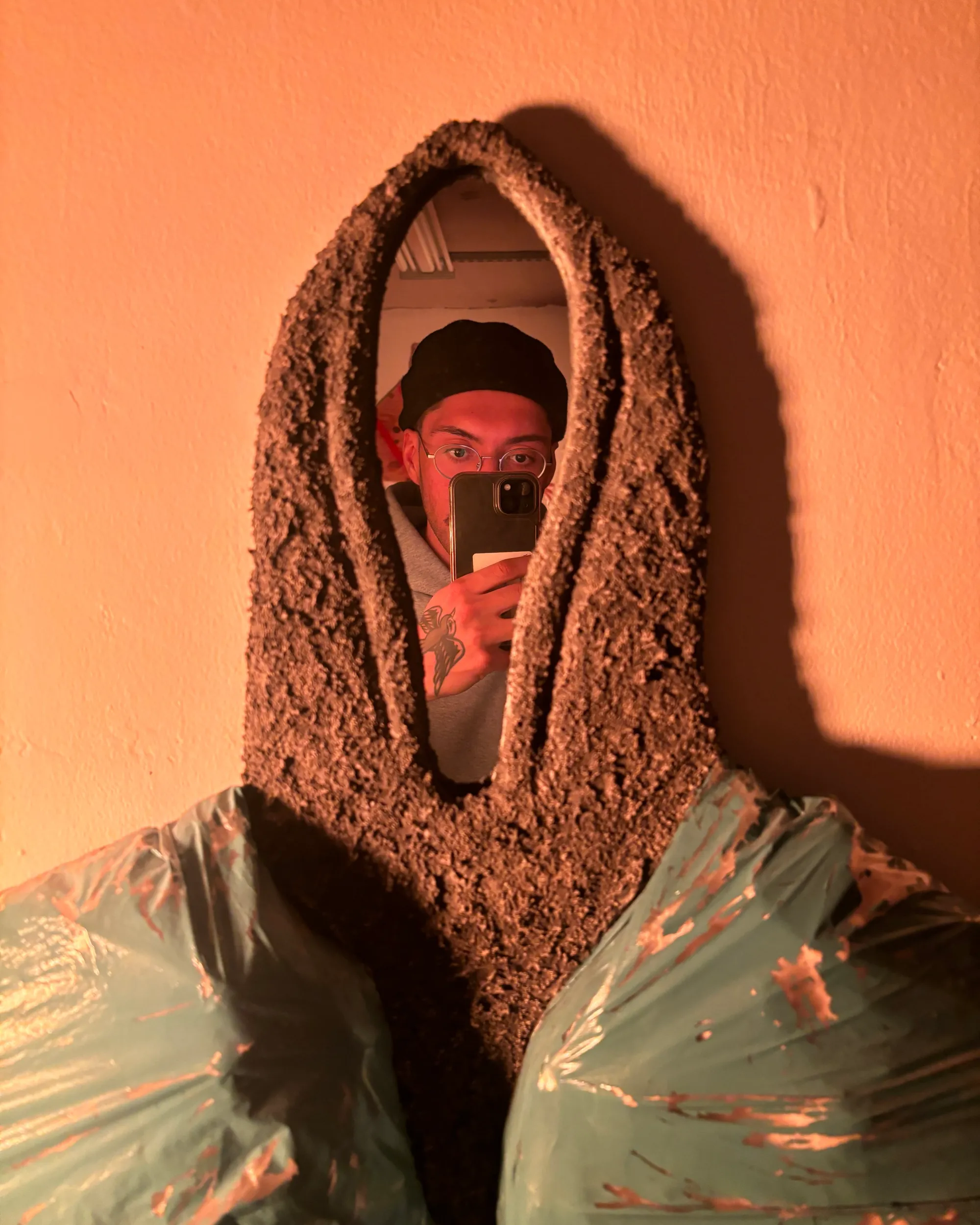 Mirror-faced hoodie reflecting the artist; textured hood with inflated bag-like shoulders; surveillance and self-portrait.