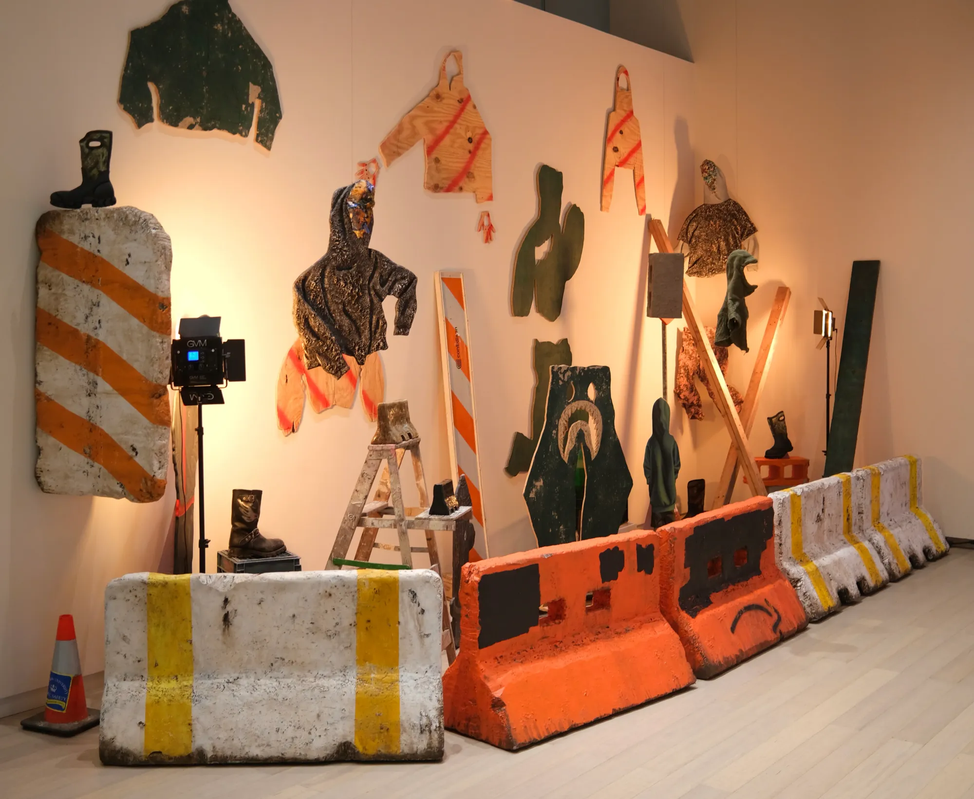 Row of orange-white concrete barriers, hanging hoodie shapes, and signage forms; construction-site aesthetics.