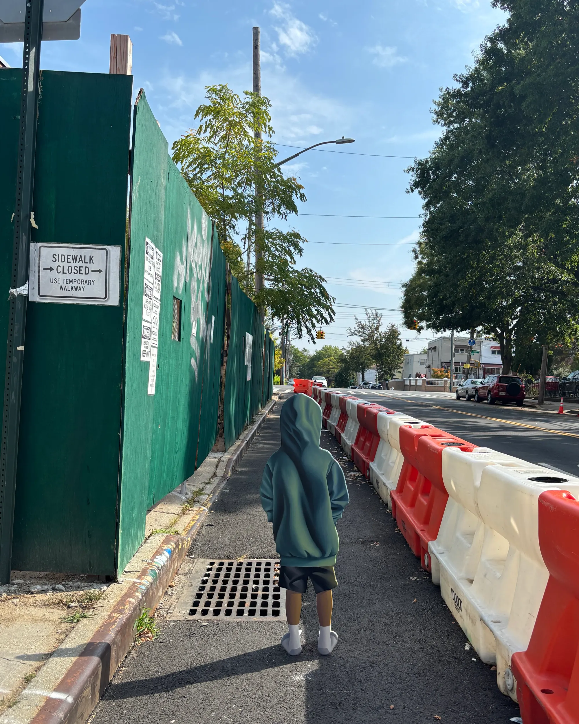 Daniel Castro street view with hooded-figure sculpture on sidewalk beside red-white construction barriers and green site fence.