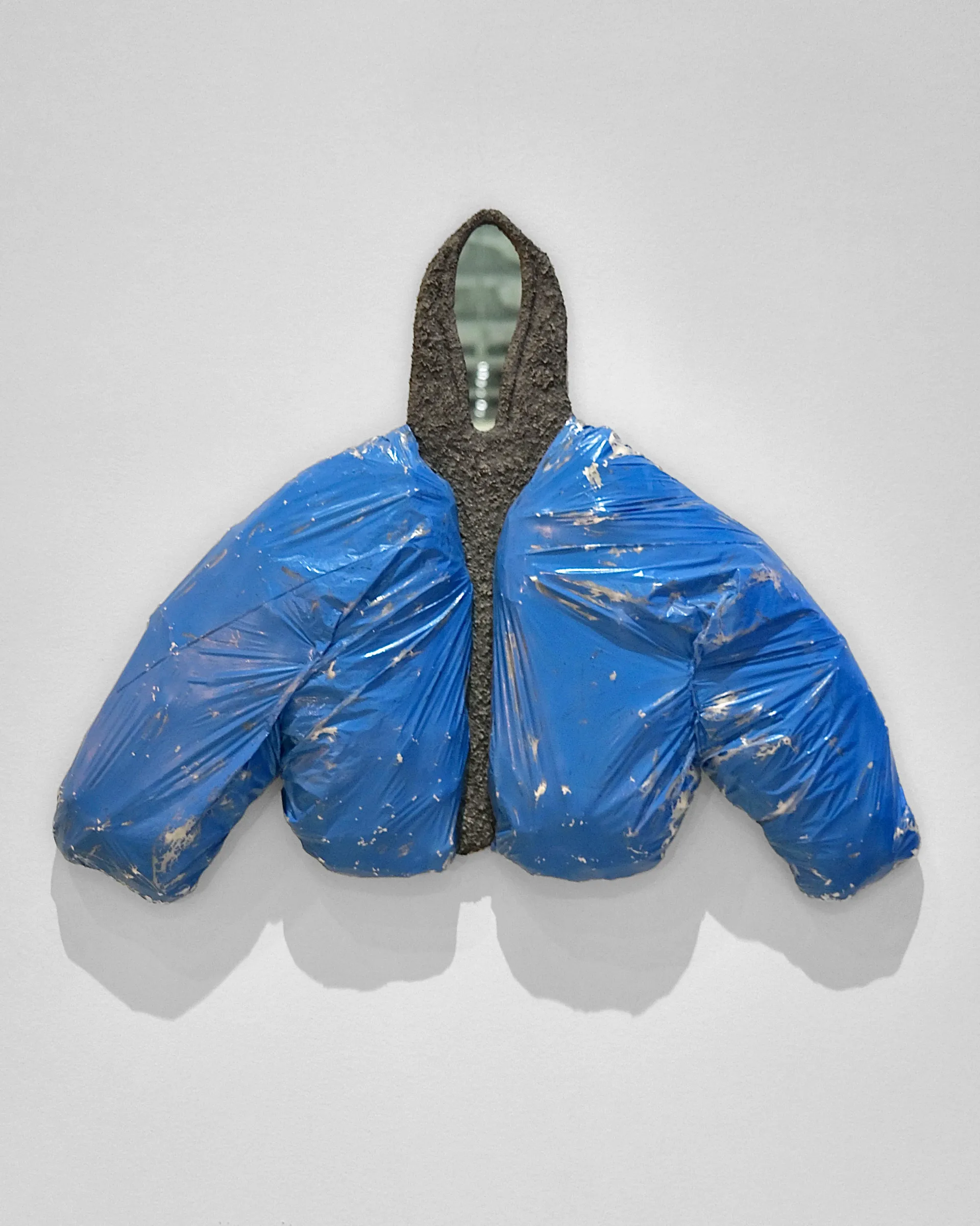 Wall-mounted blue puffer jacket with a mirror in the hood; clothing as sculpture, protection and scrutiny.
