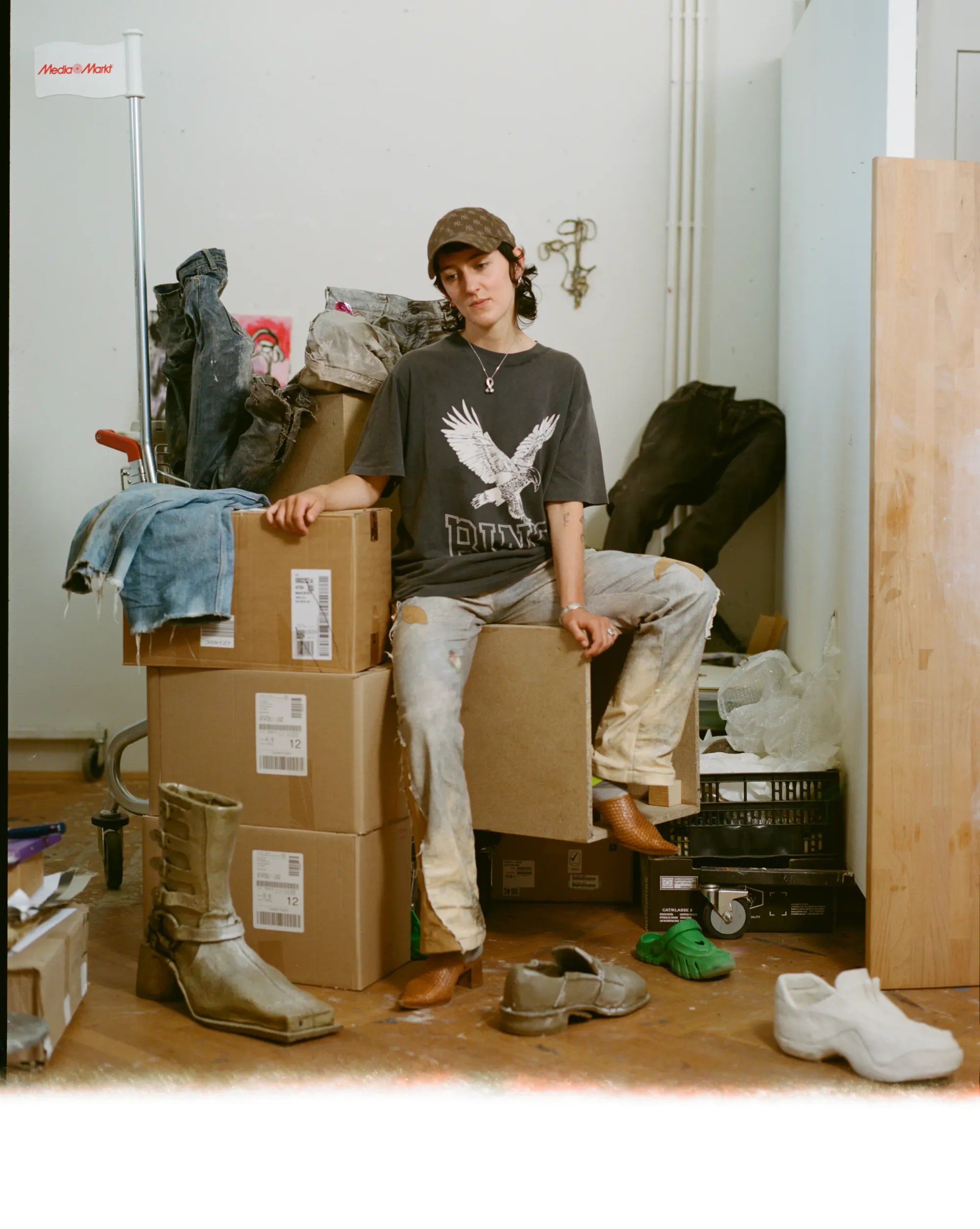 Ju Aichinger seated on their own artwork—artist portrait in a studio/gallery, black turtleneck and cap, mixed shoes, textile/sculptural piece across lap, paint buckets in background; Vienna-based artist working in ceramics, costume, and performance.