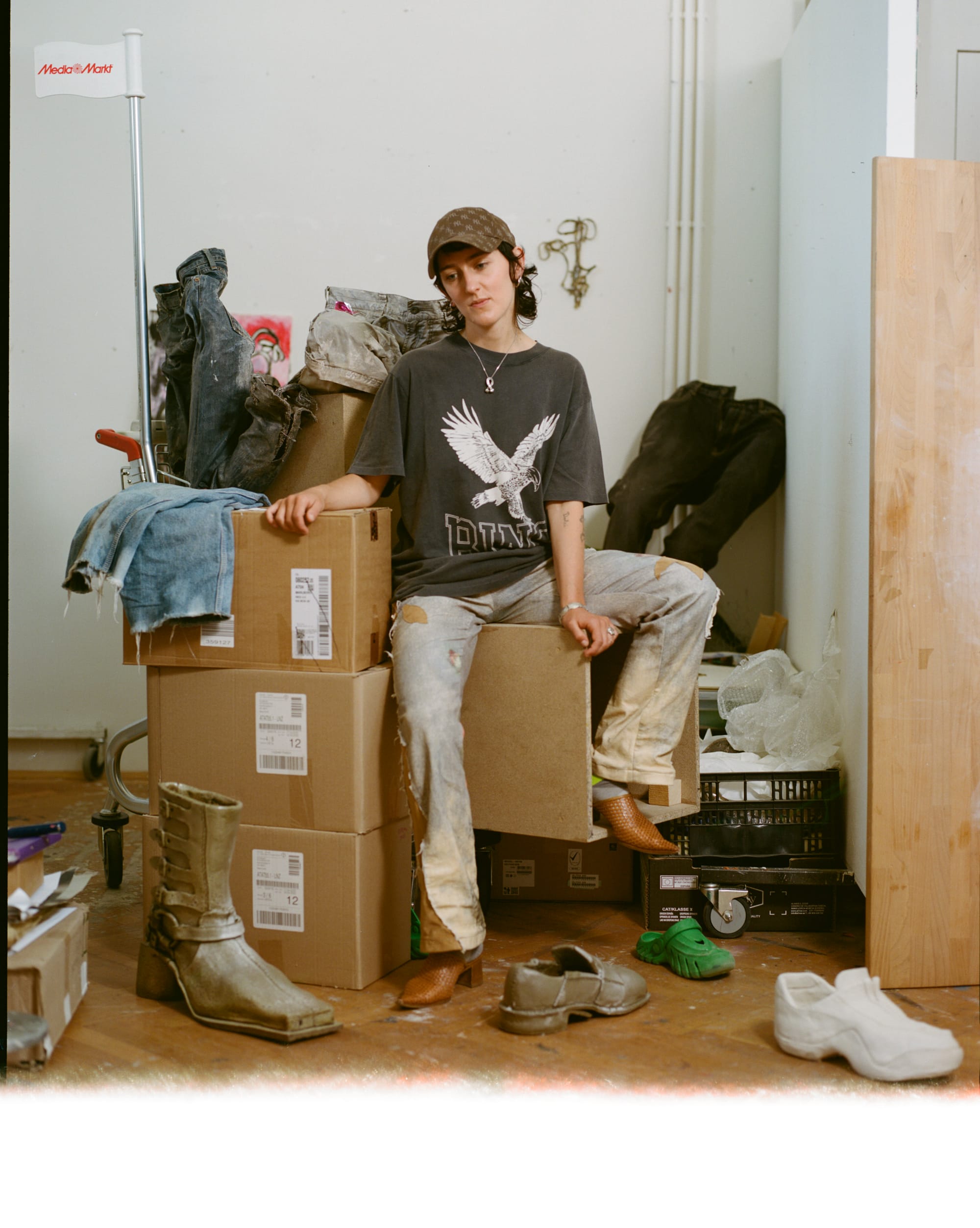 Portrait of Austrian artist Ju Aichinger, seated in a minimalist space wearing black and green footwear, holding fabric and sculptural materials. Aichinger, known for transforming everyday objects into conceptual works exploring queerness, intimacy, and social presence, will present an Artist Statement at Parallel Vienna 2025, a leading alternative art fair showcasing emerging and contemporary talents.