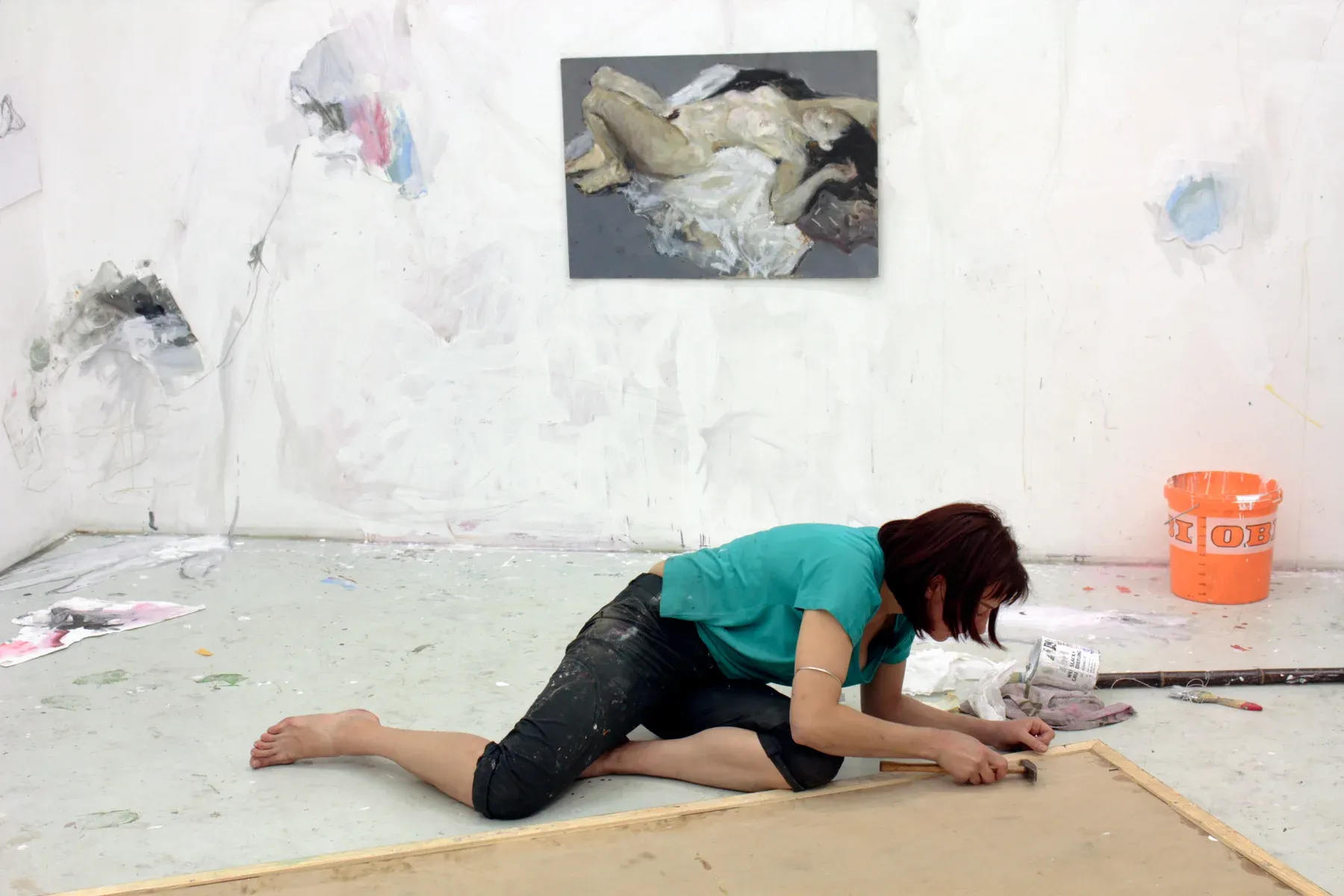Maria Naidyonova working on the floor of her studio, stretching or preparing a large canvas, surrounded by tools and unfinished figurative works on the walls.