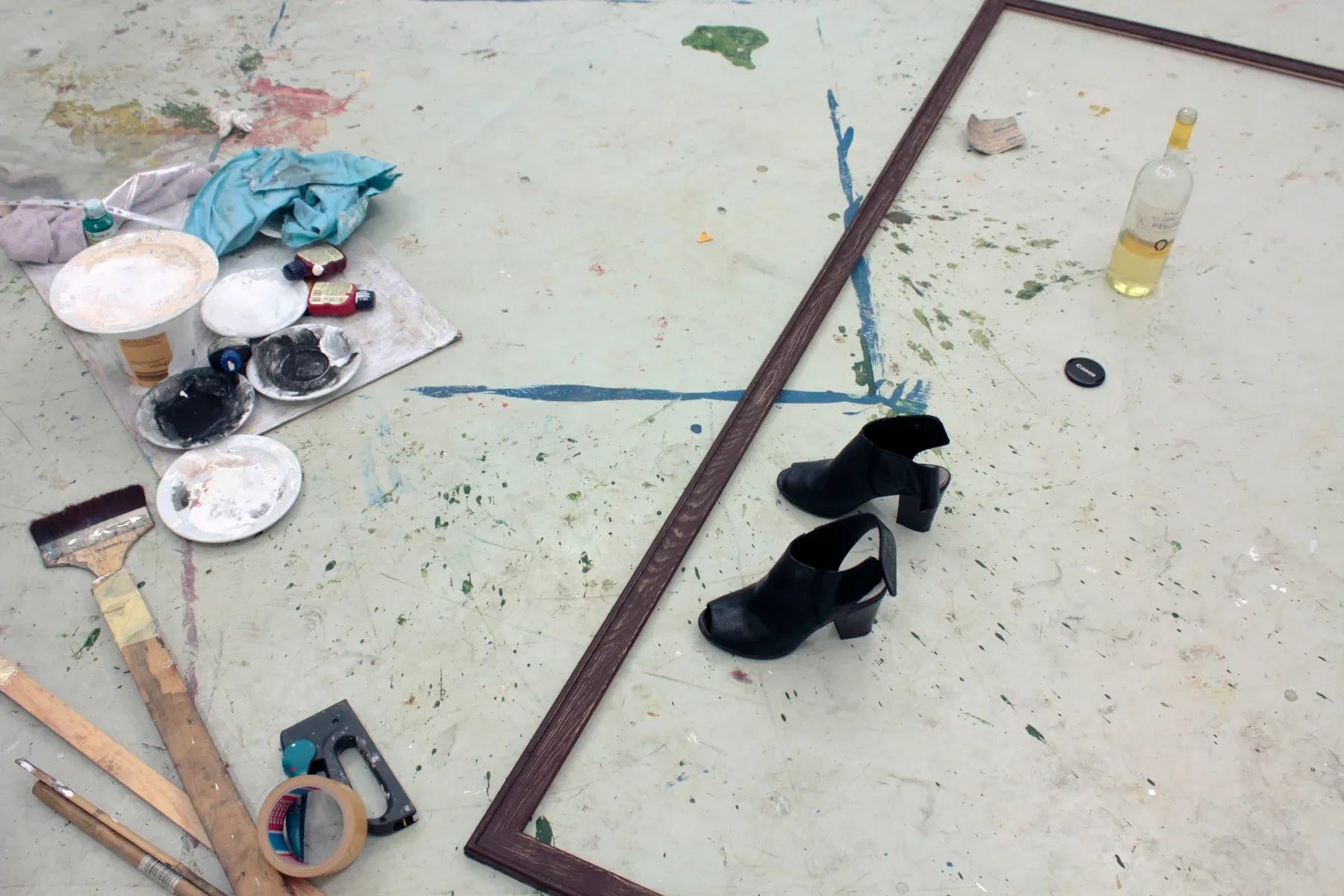 An overhead studio floor scene showing Maria Naidyonova’s tools, paint containers, black high-heeled shoes, a frame, brushes, and a bottle, indicating an active working process.