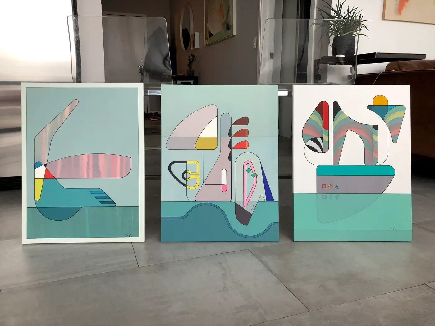 Three abstract geometric paintings by Chris Cassimatis displayed on a floor in Chicago, each with layered shapes, pastel tones and structural linework. Image Courtesy of the Artist.