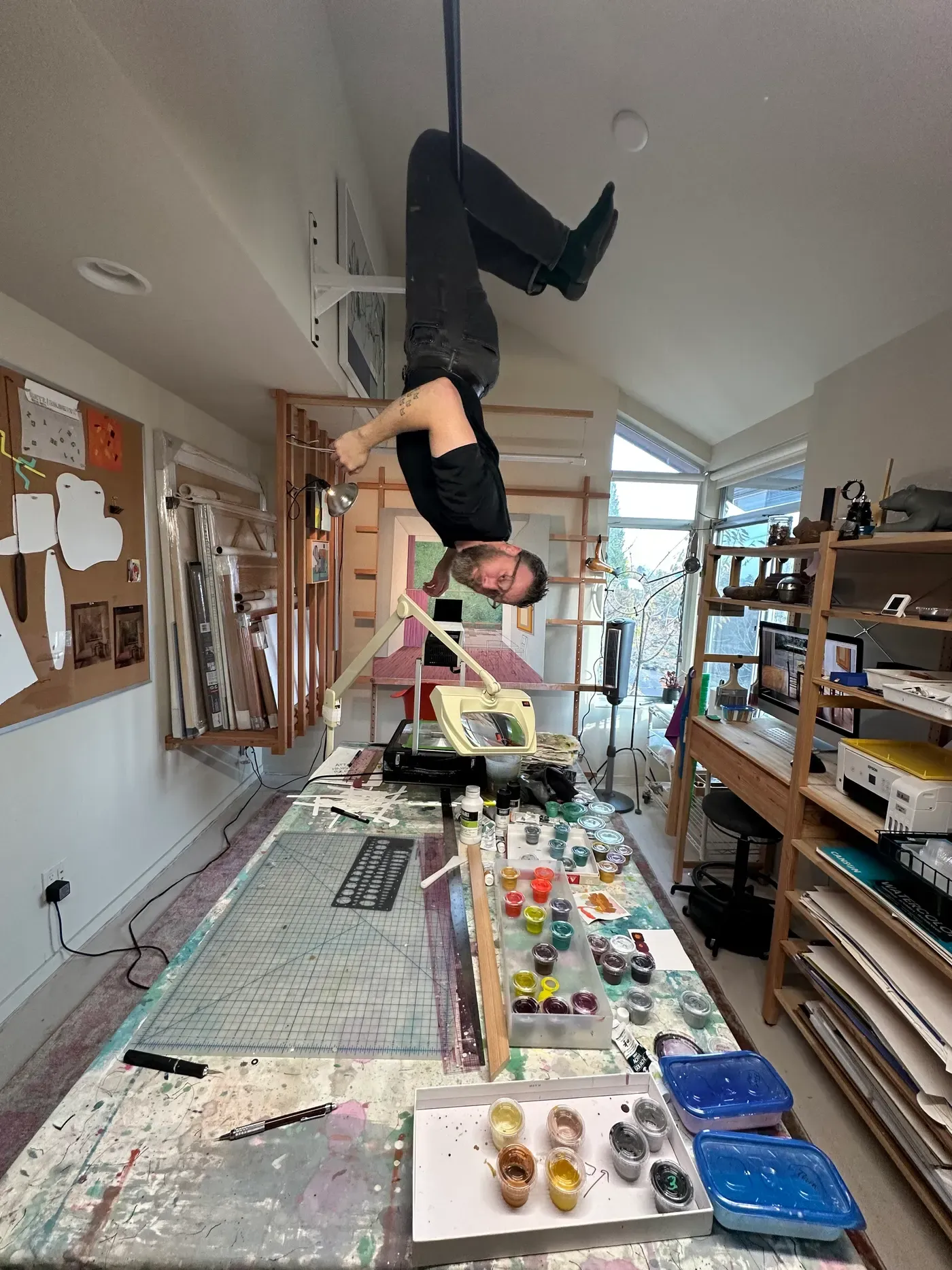 Artist Chris Cassimatis hanging upside down in his studio above a large work table covered with inks, tools and drawings, showing his playful approach to art making.