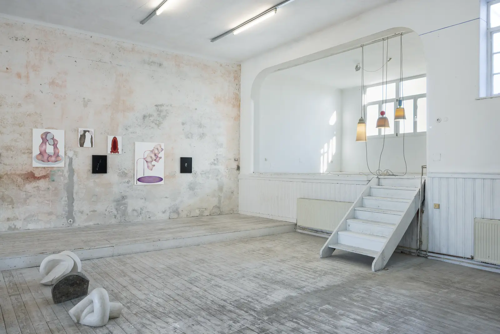 Exhibition view at Kunstverein Schattendorf showing paintings on a textured wall, a white staircase, hanging lamps and sculptural works placed on the wooden floor.
