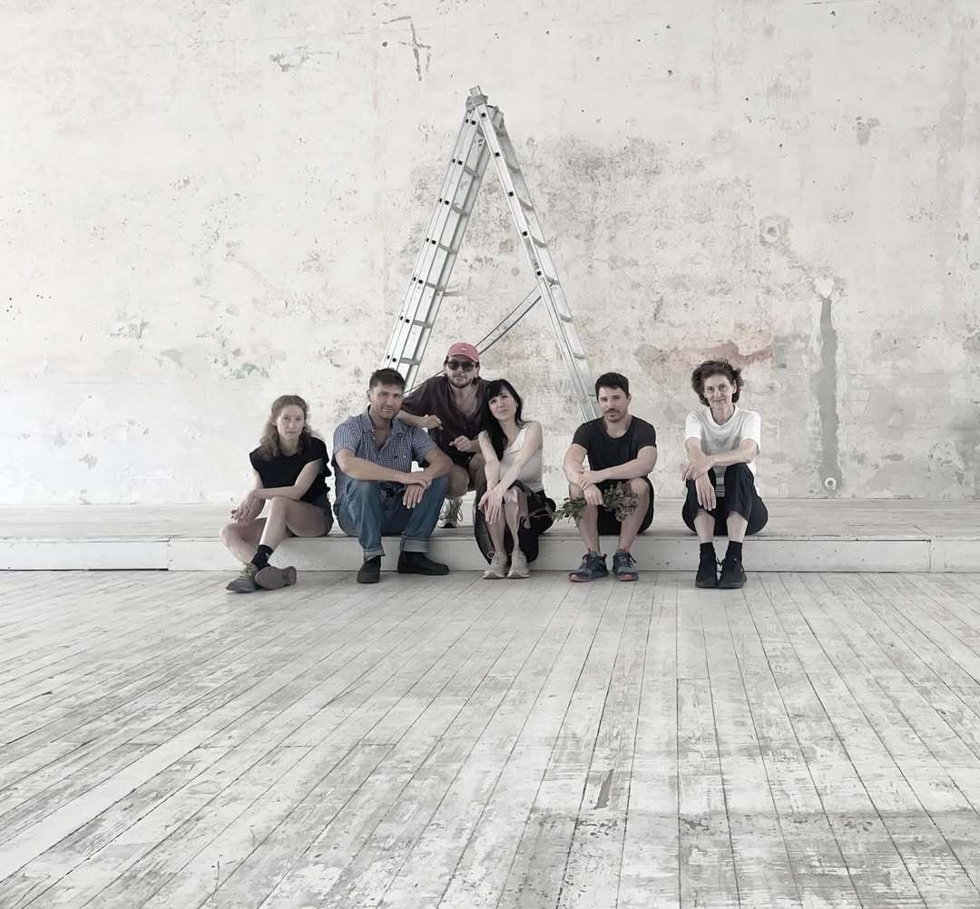 Group photo of the Alaska artists sitting on the wooden floor at Kunstverein Schattendorf, with a ladder behind them and a textured wall in the background.