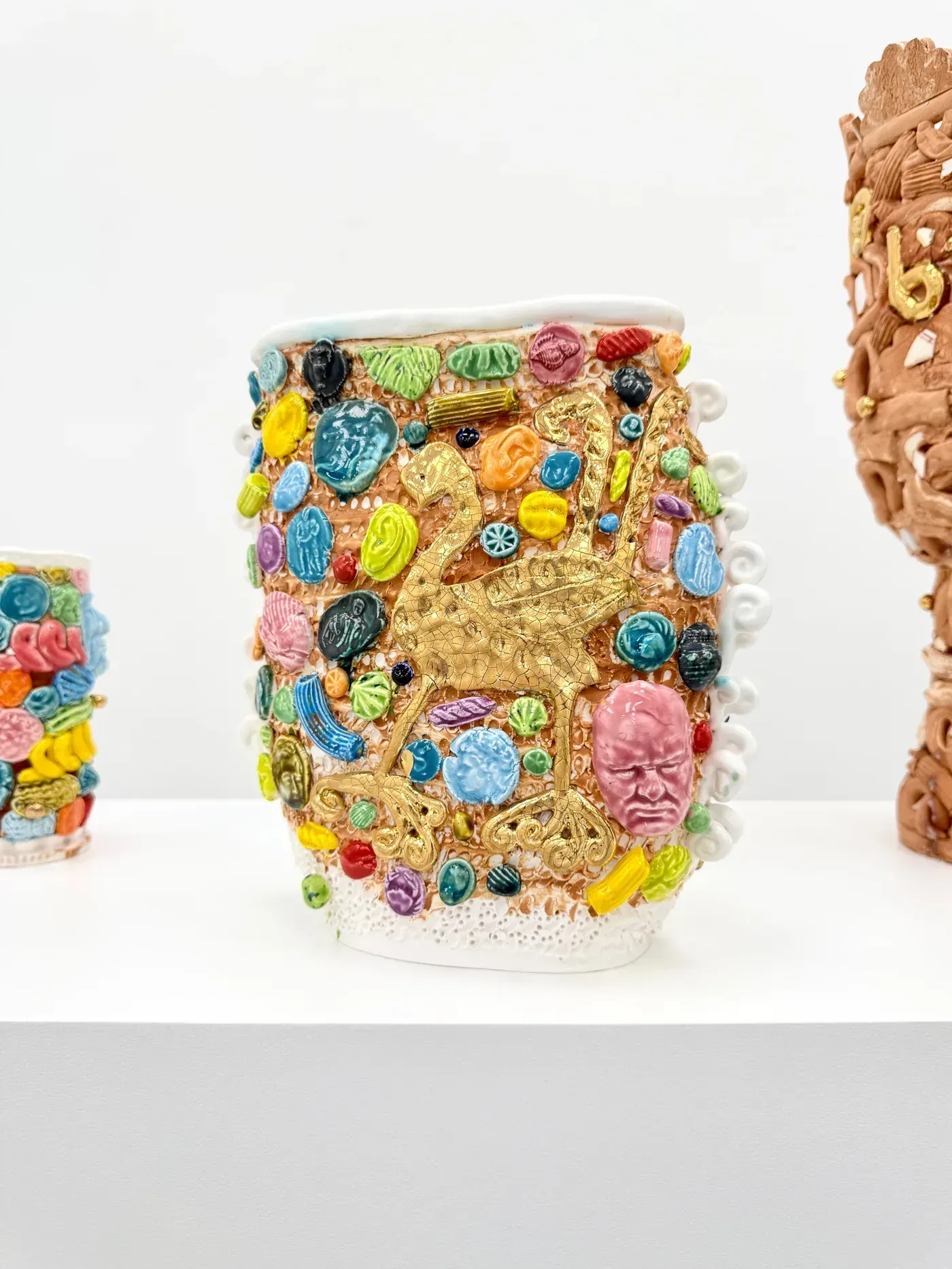 new ceramic artists now: A richly decorated ceramic vase by Glenn Barkley covered in colorful glazed ornaments, small cameo faces, and a gold lyrebird motif, displayed on a white pedestal at HAGD Contemporary in Aalbor