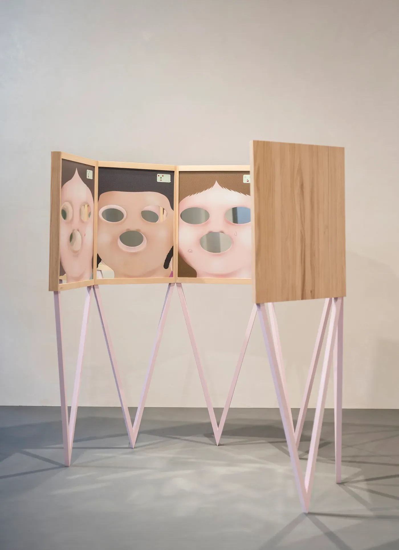 Sculptural installation Memoràndum del Quotidià by Grip Face. Three painted wooden panels on pink angular legs show cartoon-like faces with hollow eyes and mouths, suggesting fragmented memory and distorted perception. Exhibited at La Misericordia in Palma with Miju Lee, curated by Sofia Moisés.