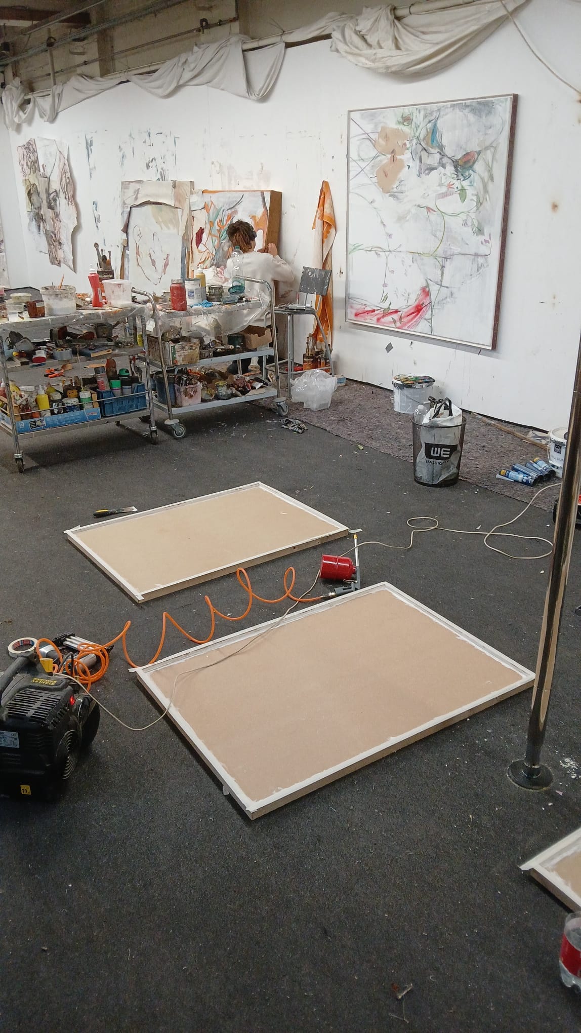 Ruth Devriendt’s studio view from 2025, with a person working on a canvas beside painting carts, tools and large works in progress. Materials and frames are scattered across the floor. Image courtesy of the artist.