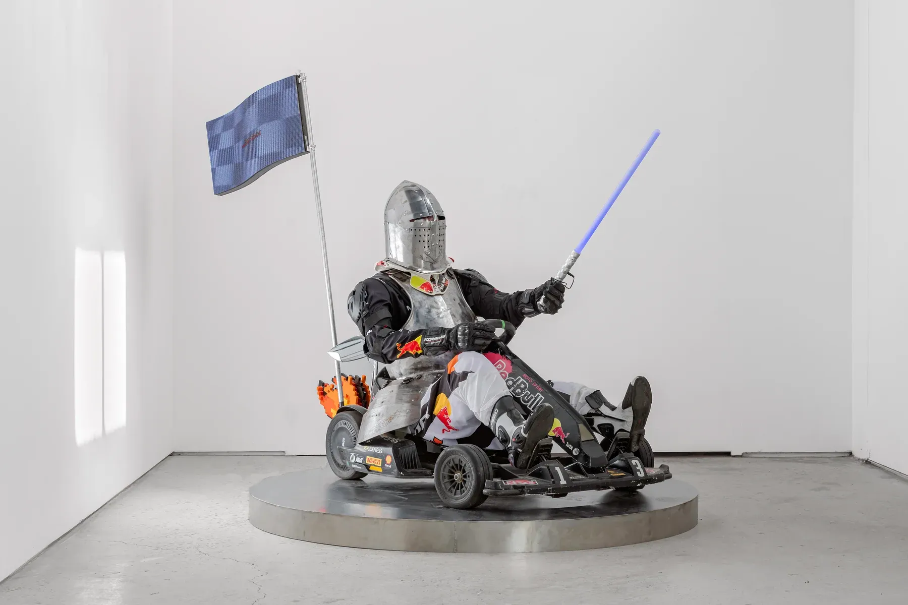 Knight like figure in armor riding a modified go kart with a raised flag, displayed on a circular platform in a white gallery.