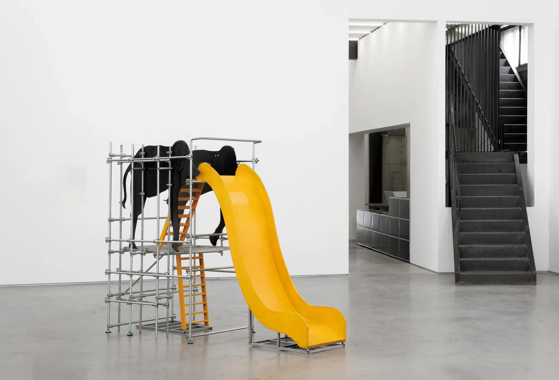 Large installation with a yellow slide attached to a metal scaffold supporting black sculptural elements, displayed in a gallery space.