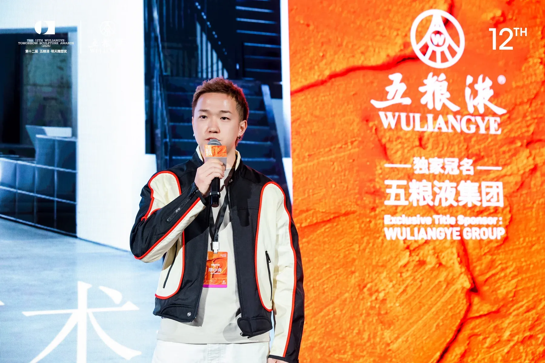 Artist Xiaozhou Liao speaking on stage at the 12th Wuliangye Tomorrow Sculpture Awards, standing in front of large event screens with the sponsor logo visible.