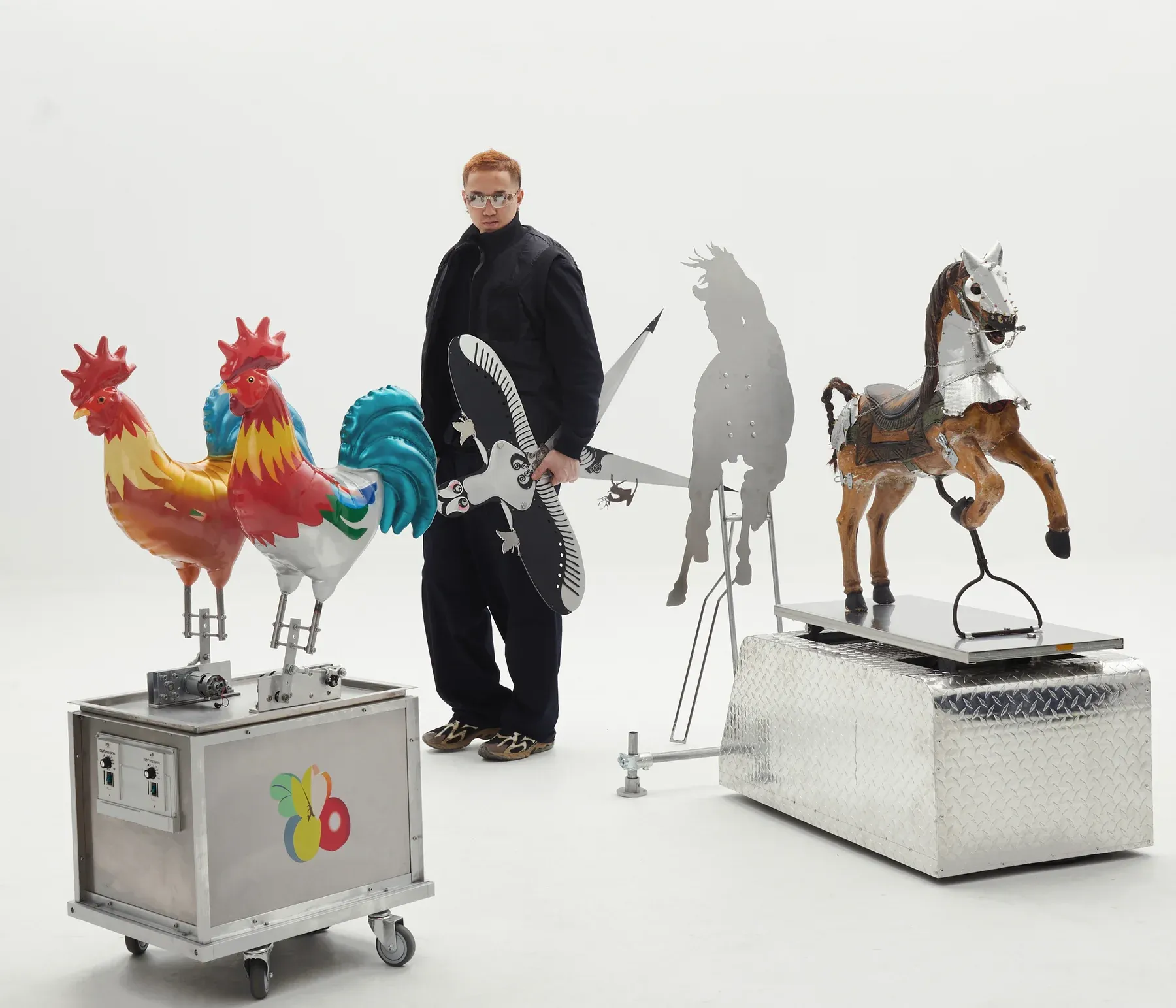 Artist Xiaozhou Liao standing among kinetic rooster sculptures and metal cutout figures in a white studio setup.