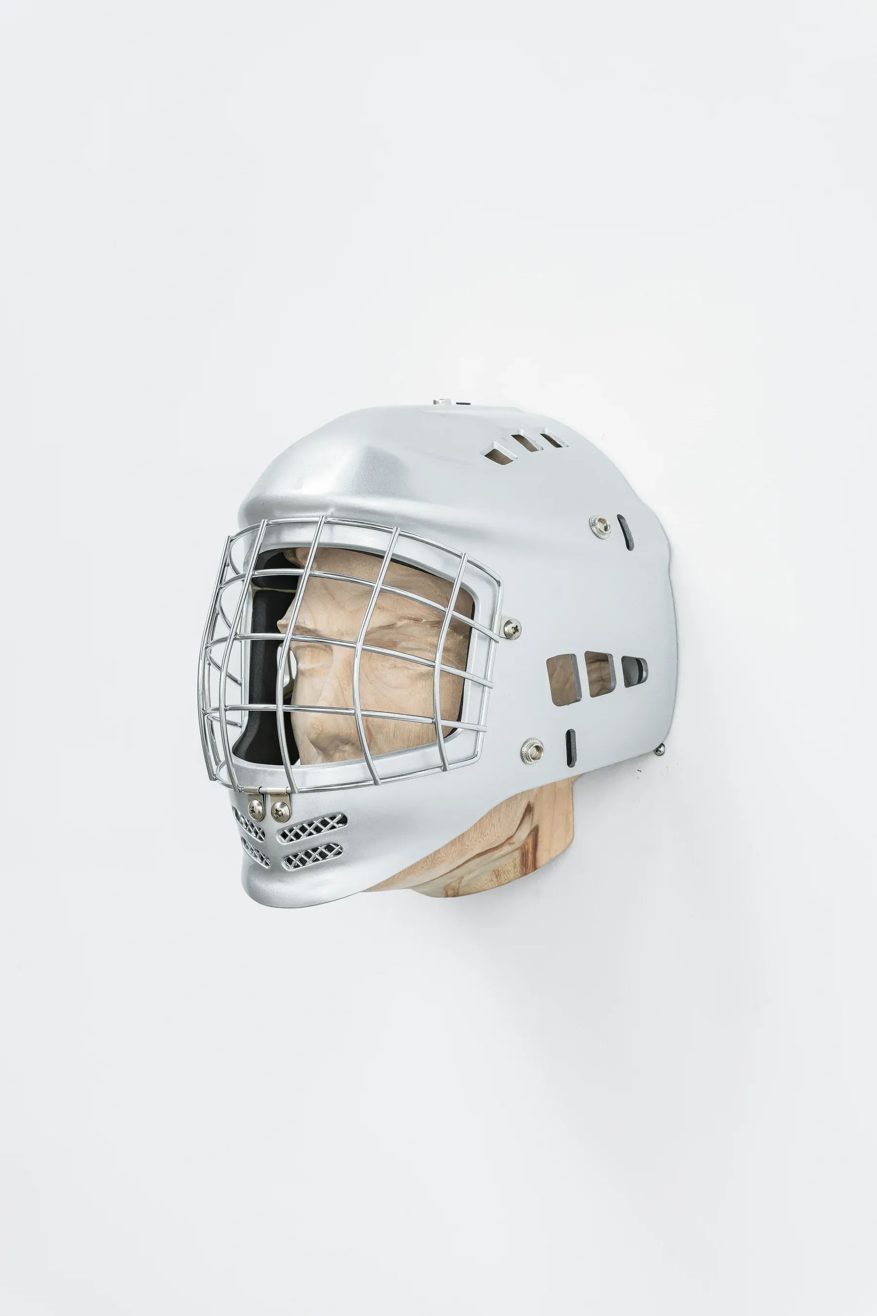 Silver helmet sculpture with a cage style face guard mounted on a wooden support against a white wall.
