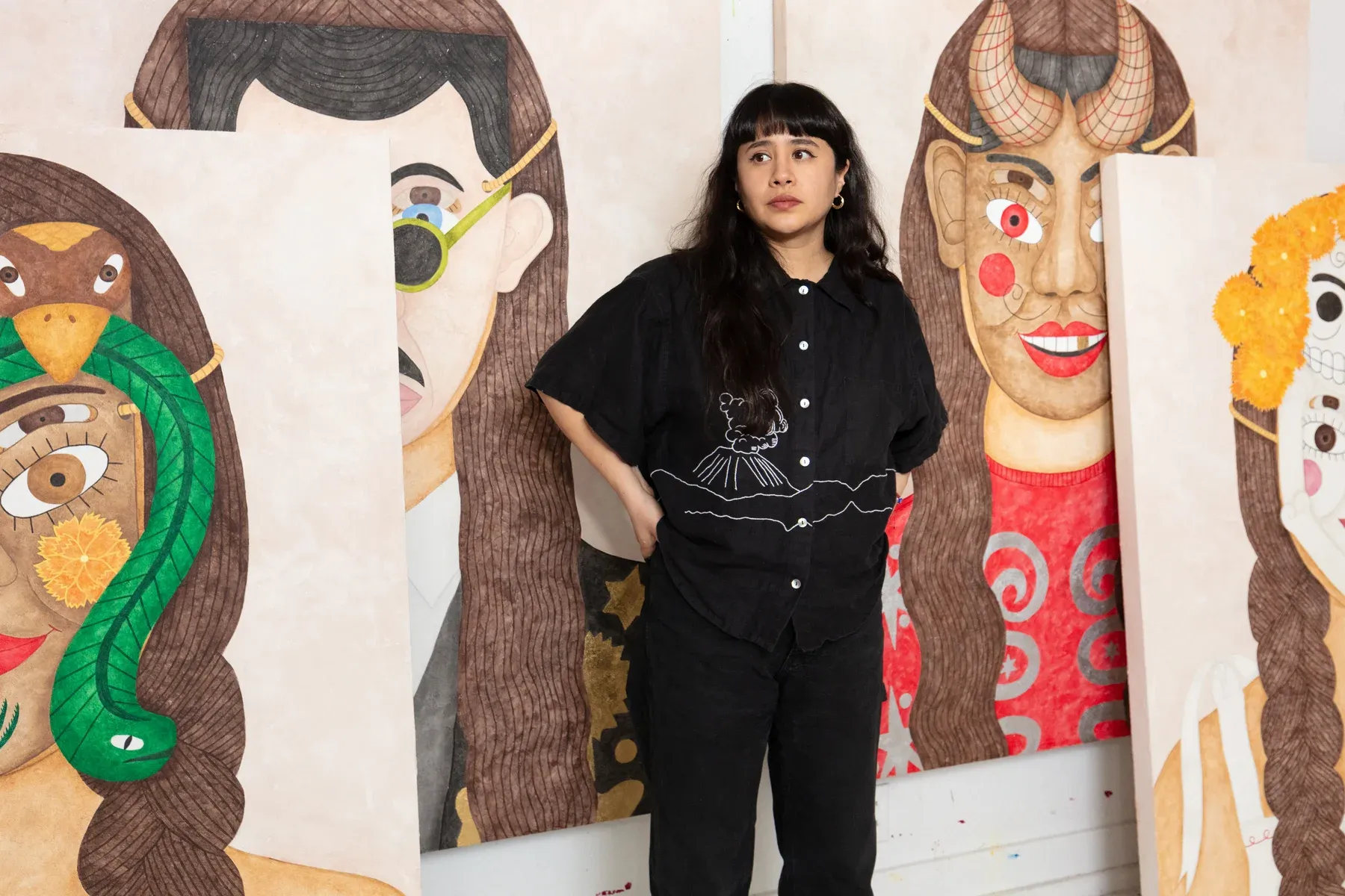 Liz Hernandez, artist standing in a studio surrounded by large painted portraits featuring mask-like composite faces.