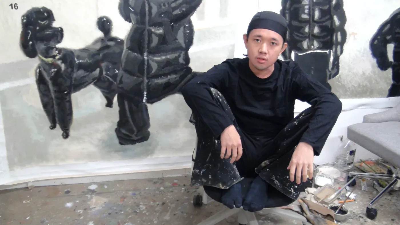 Yutaro Inagaki seated in his studio in front of large black puffer coated figure paintings.