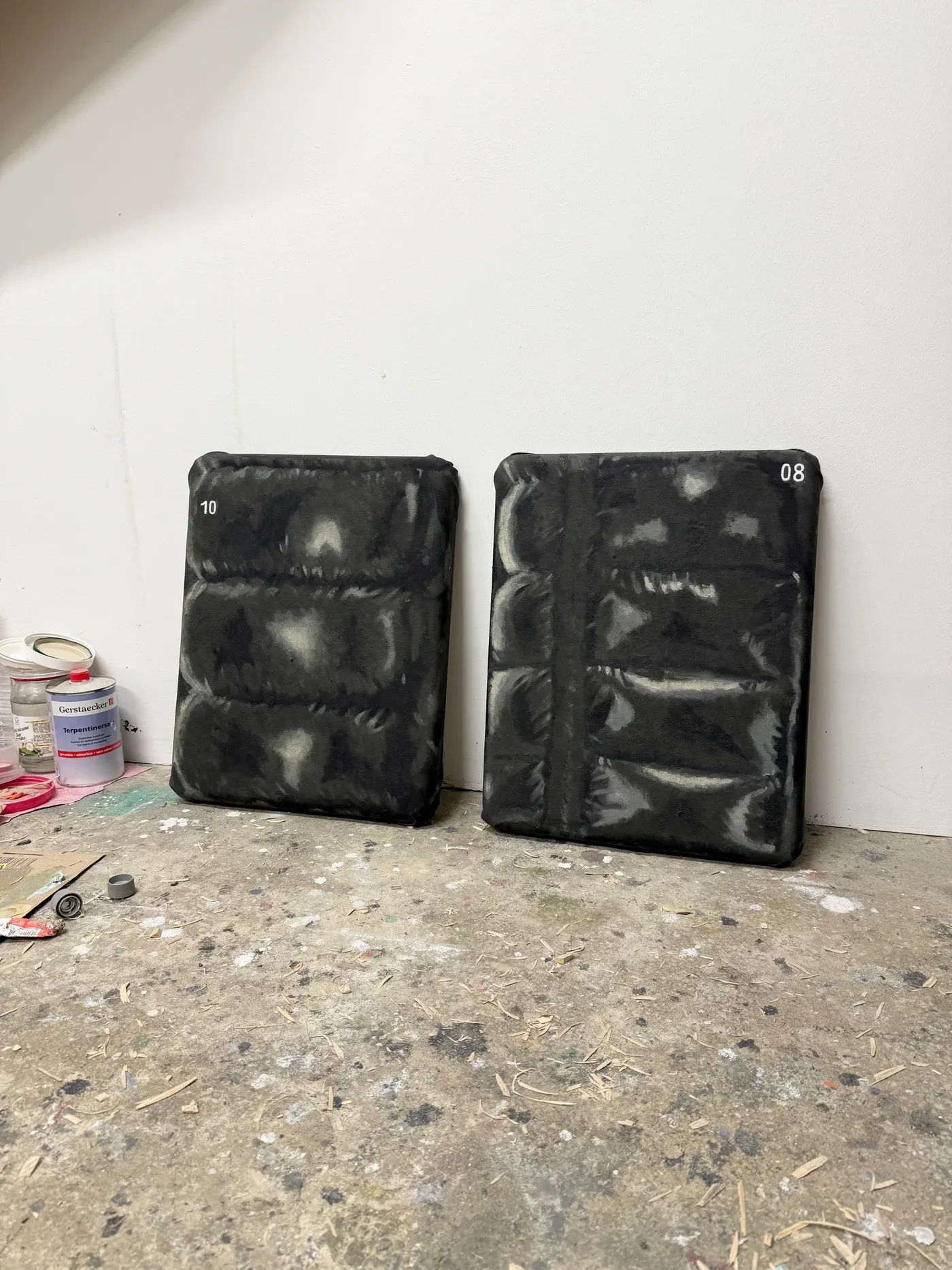 artist pages: Yutaro Inagaki, Two small oil paintings showing close up padded puffer jacket surfaces, numbered 10 and 08, leaning against a studio wall on a paint spattered floor.