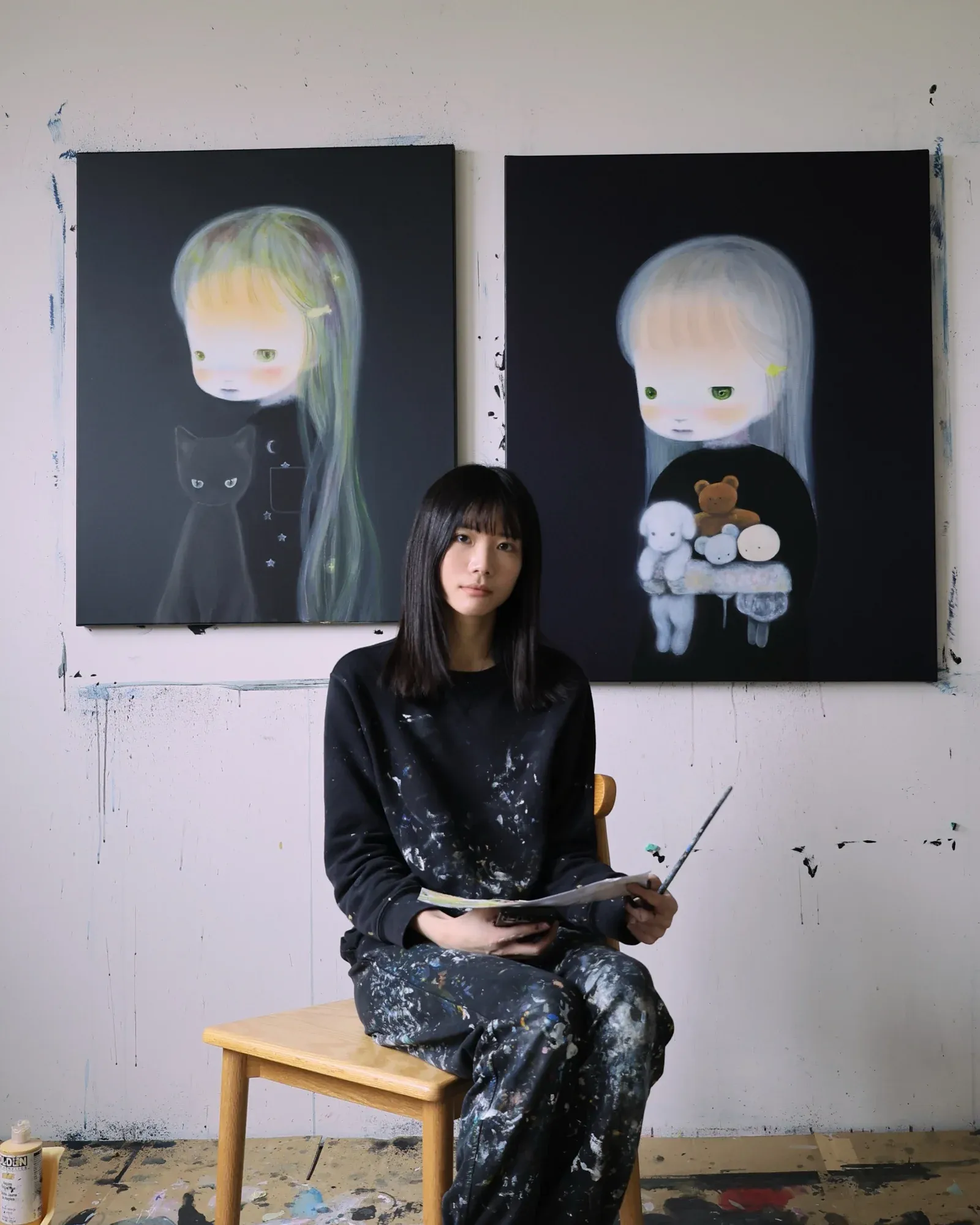Studio view of Ruo-Hsin Wu seated on a chair holding painting tools, with two finished paintings behind her showing pale figurative figures against dark backgrounds.