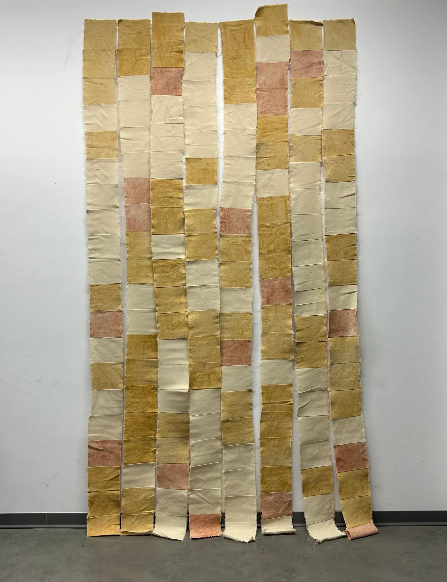 Vertical strips of canvas fabric dyed with natural pigments in muted yellow, beige and soft pink tones, stitched together to form a hanging textile surface.