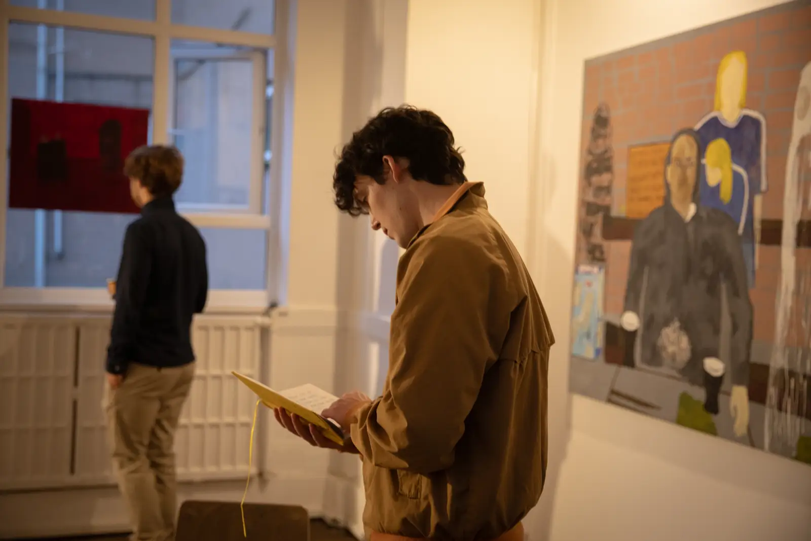 A visitor reading a yellow notebook inside Paes’ exhibition space, surrounded by figurative paintings
