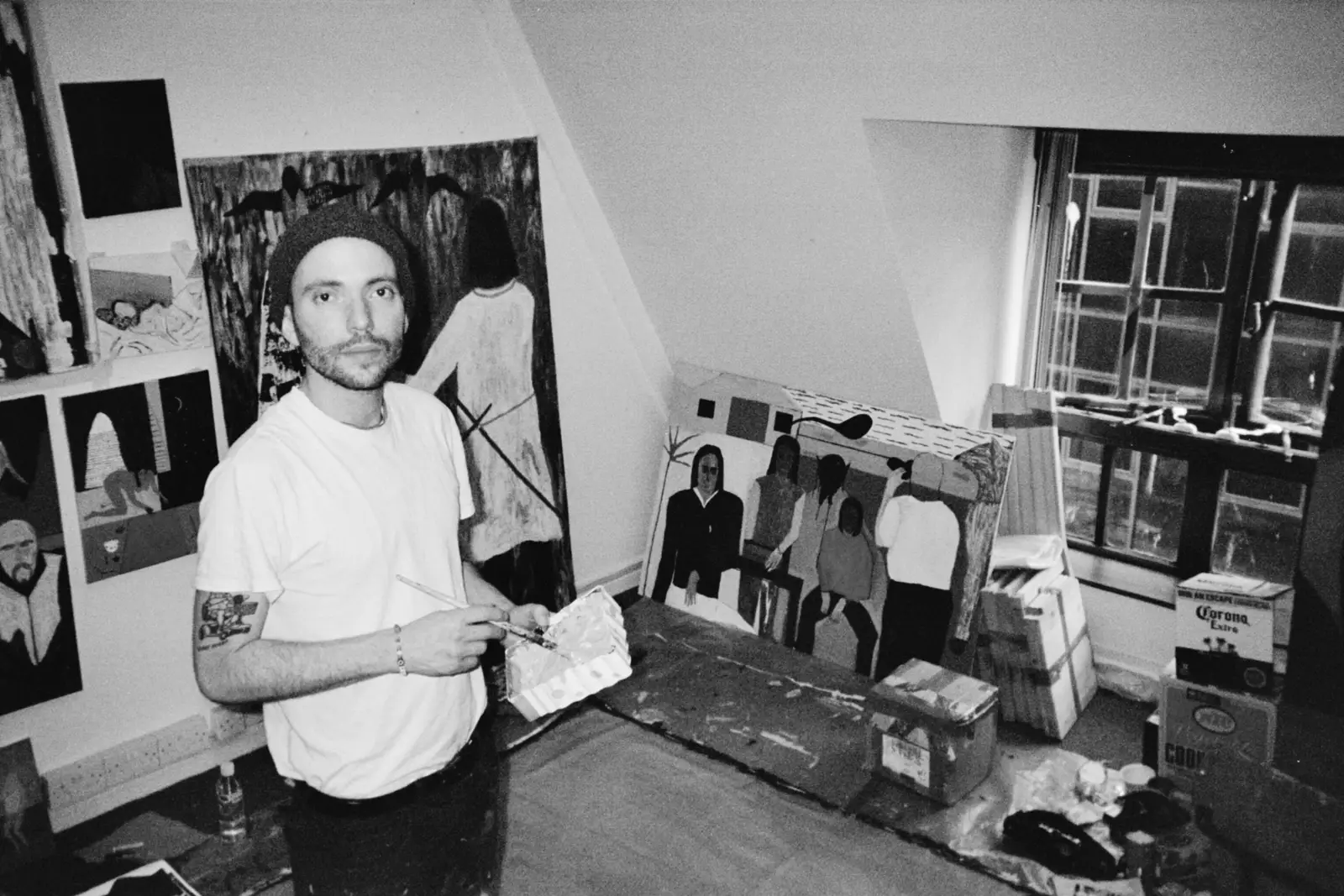 Black-and-white portrait of artist Paes in his studio holding a paint palette, surrounded by paintings and works in progress.