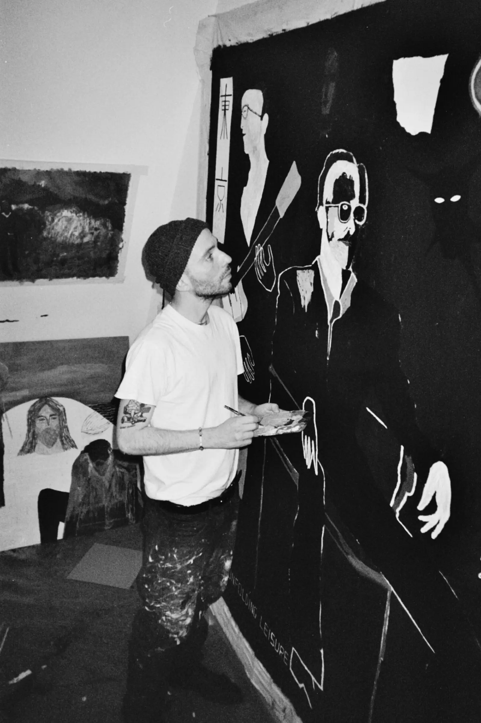 Portrait of artist Paes crouching in front of a tall painting with three figures, photographed in the studio