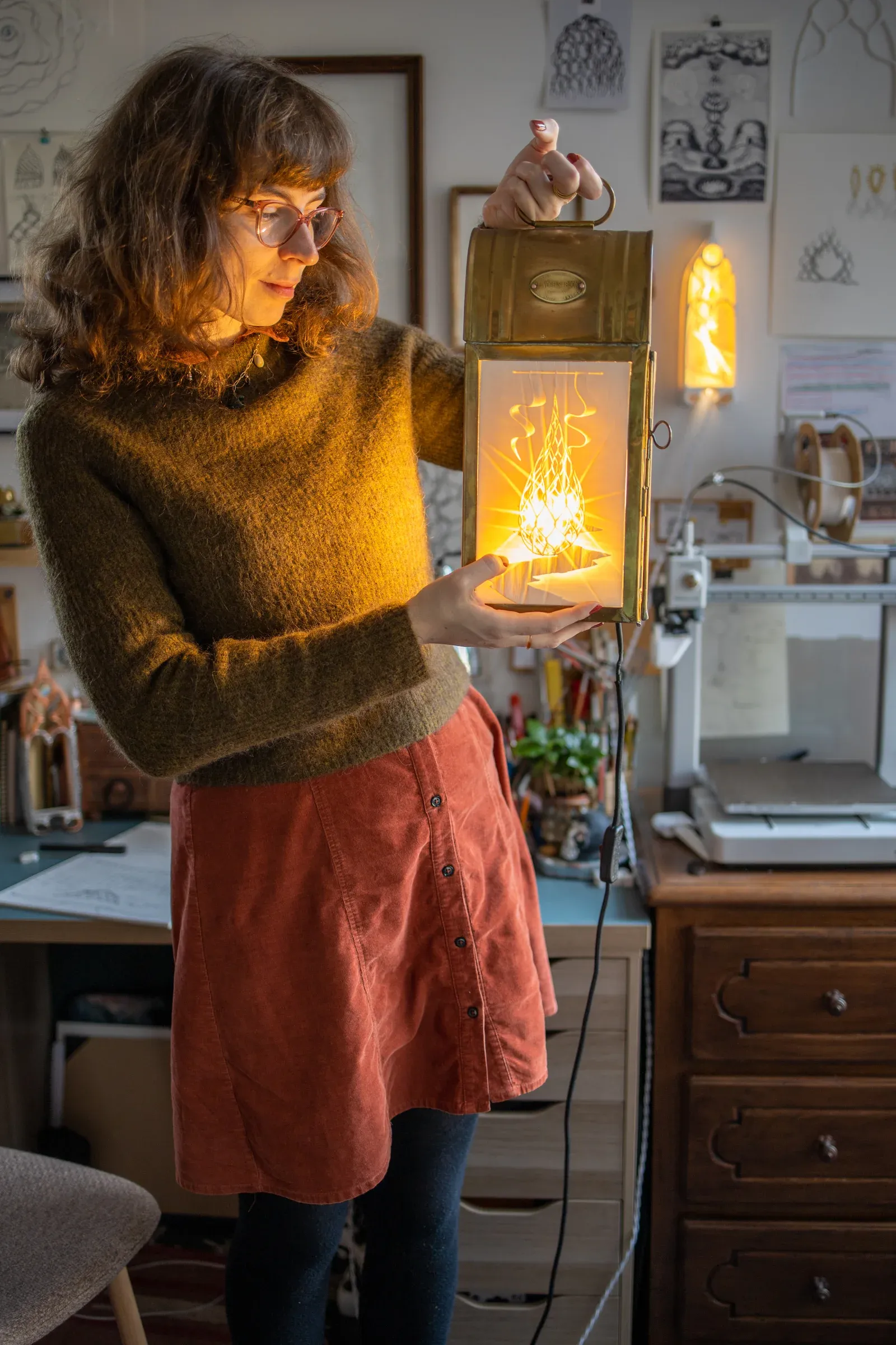Martha Kicsiny in her Ghent studio holding a glowing light-based lithophane sculpture, surrounded by drawings and historical media references.