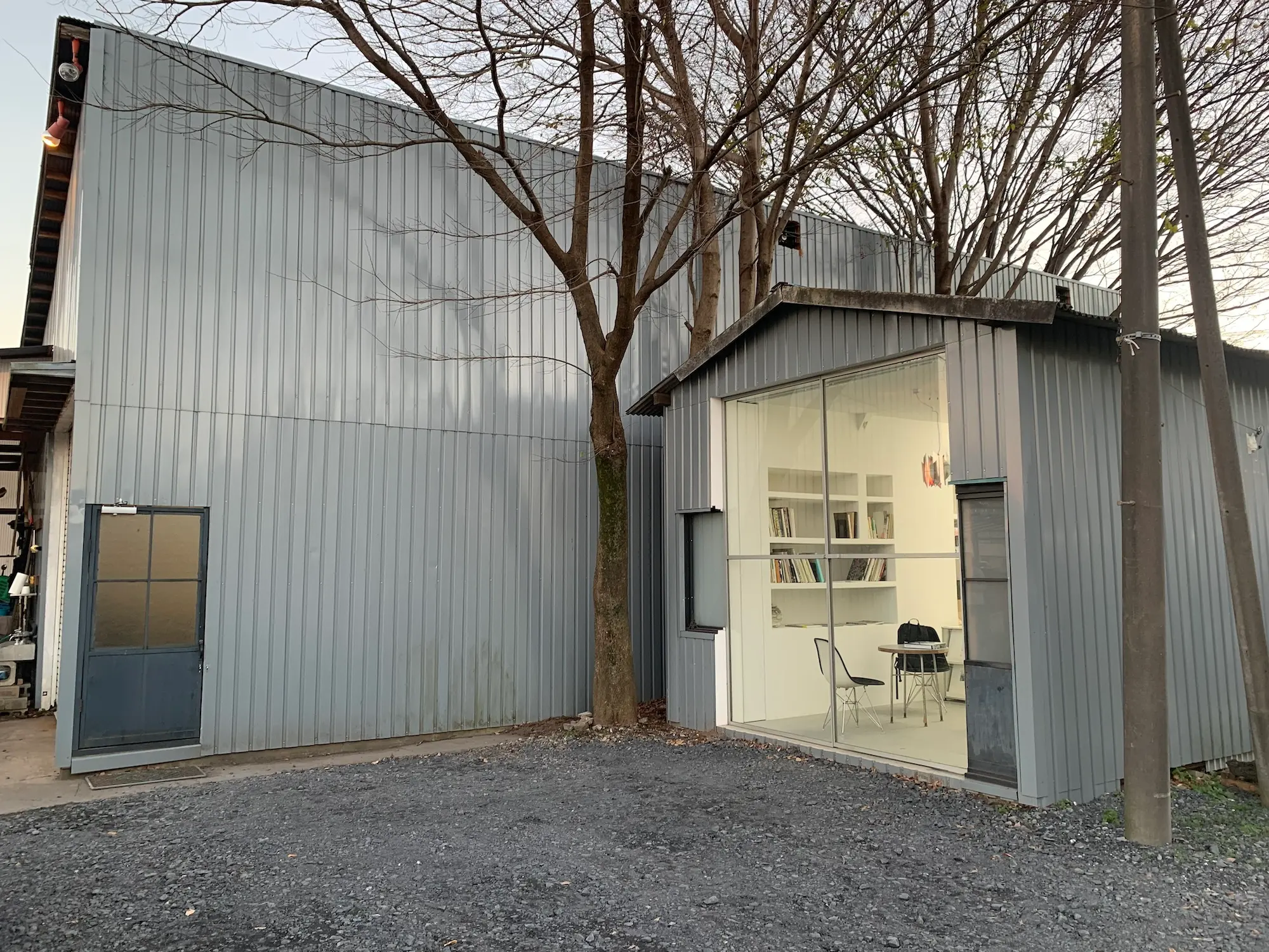 目 [mé] studio building with corrugated metal façade and a glass-fronted office extension, surrounded by trees and gravel courtyard.