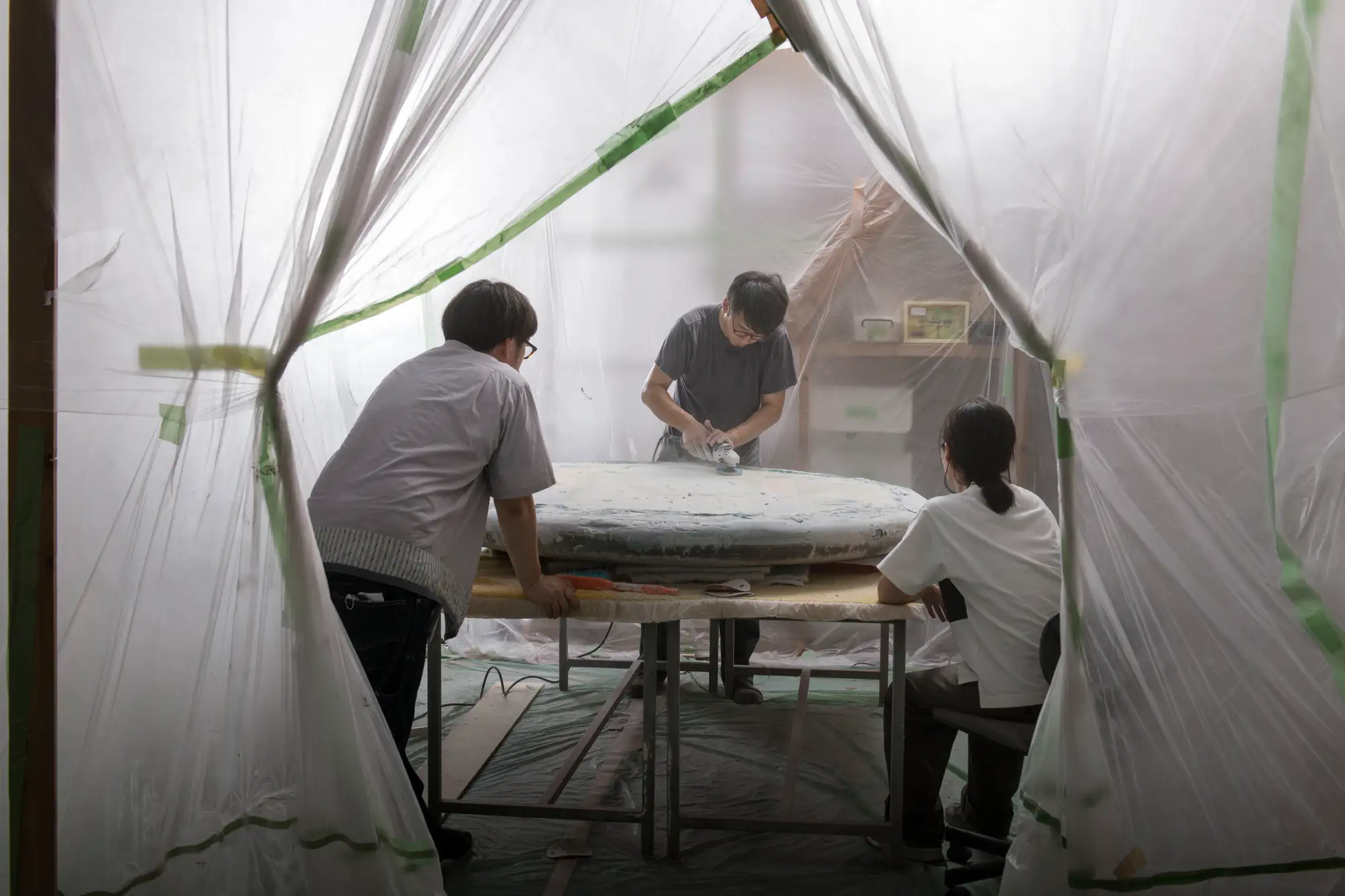 目 [mé] working inside a temporary plastic-enclosed studio environment, shaping a large circular sculptural surface during the production process.