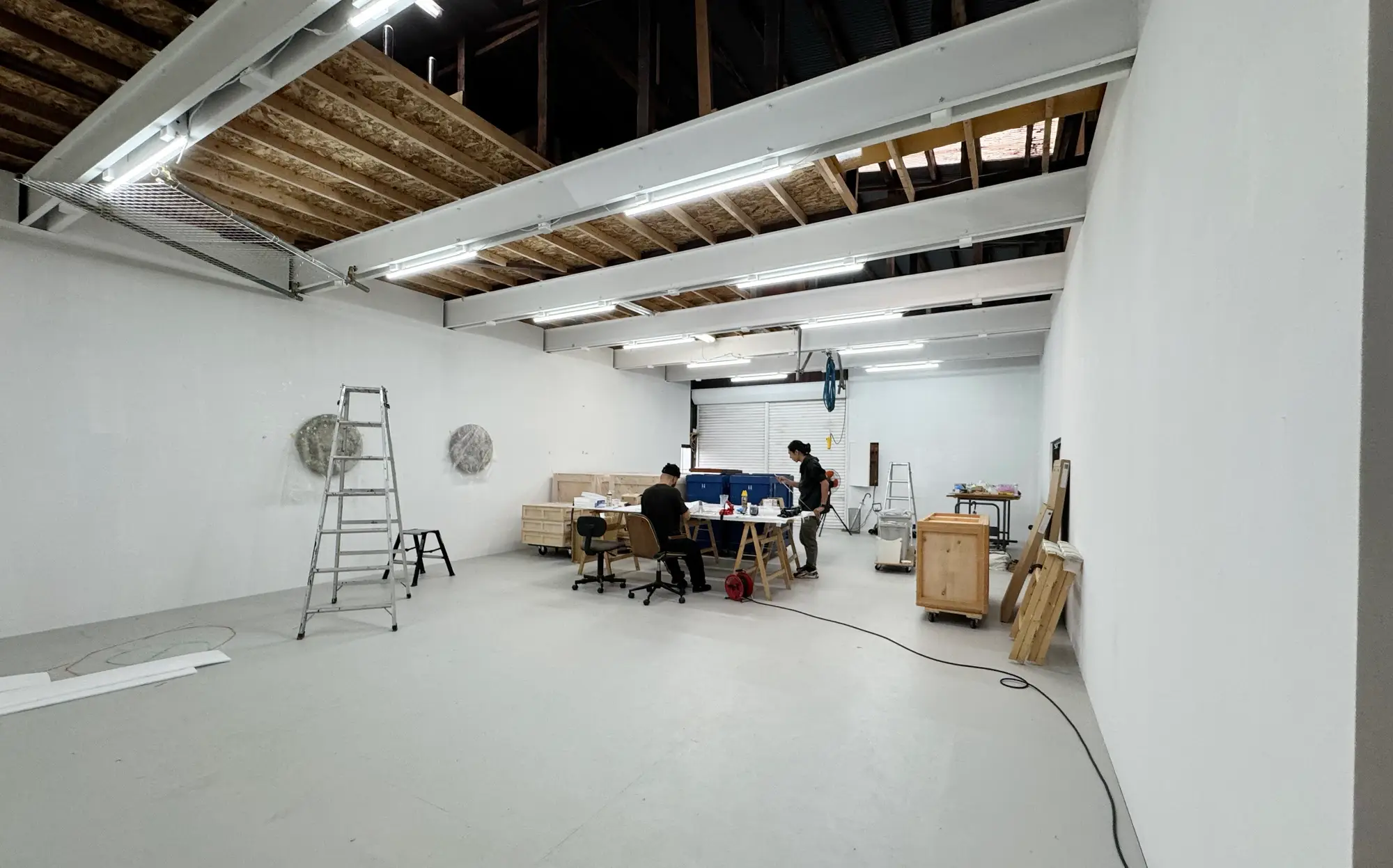 目 [mé] studio interior with white walls, exposed ceiling beams and worktables, showing an active production space with tools and sculptural elements in progress